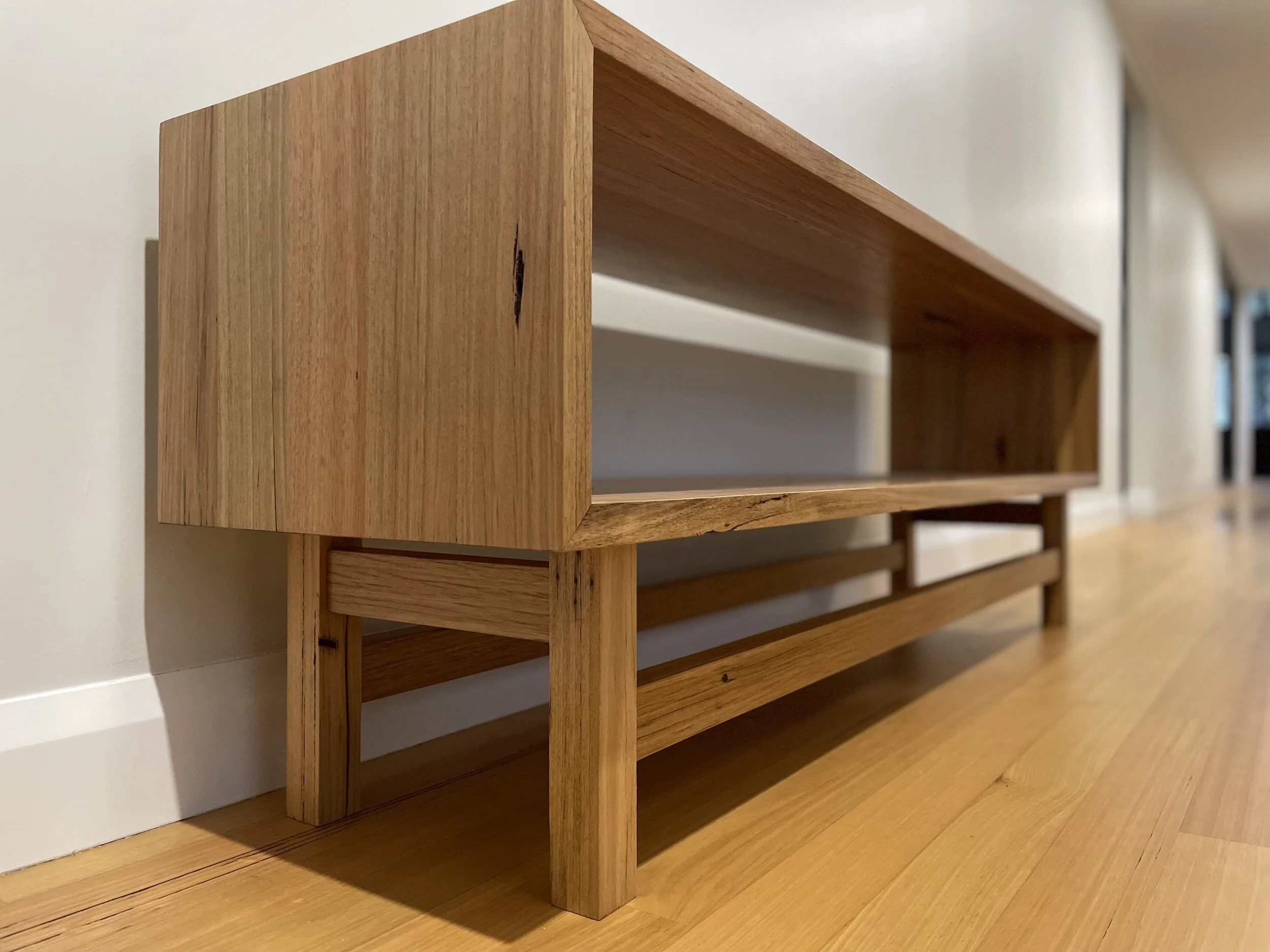 A wooden shelf with a open rectangular design, placed on a wooden floor against a white wall.
