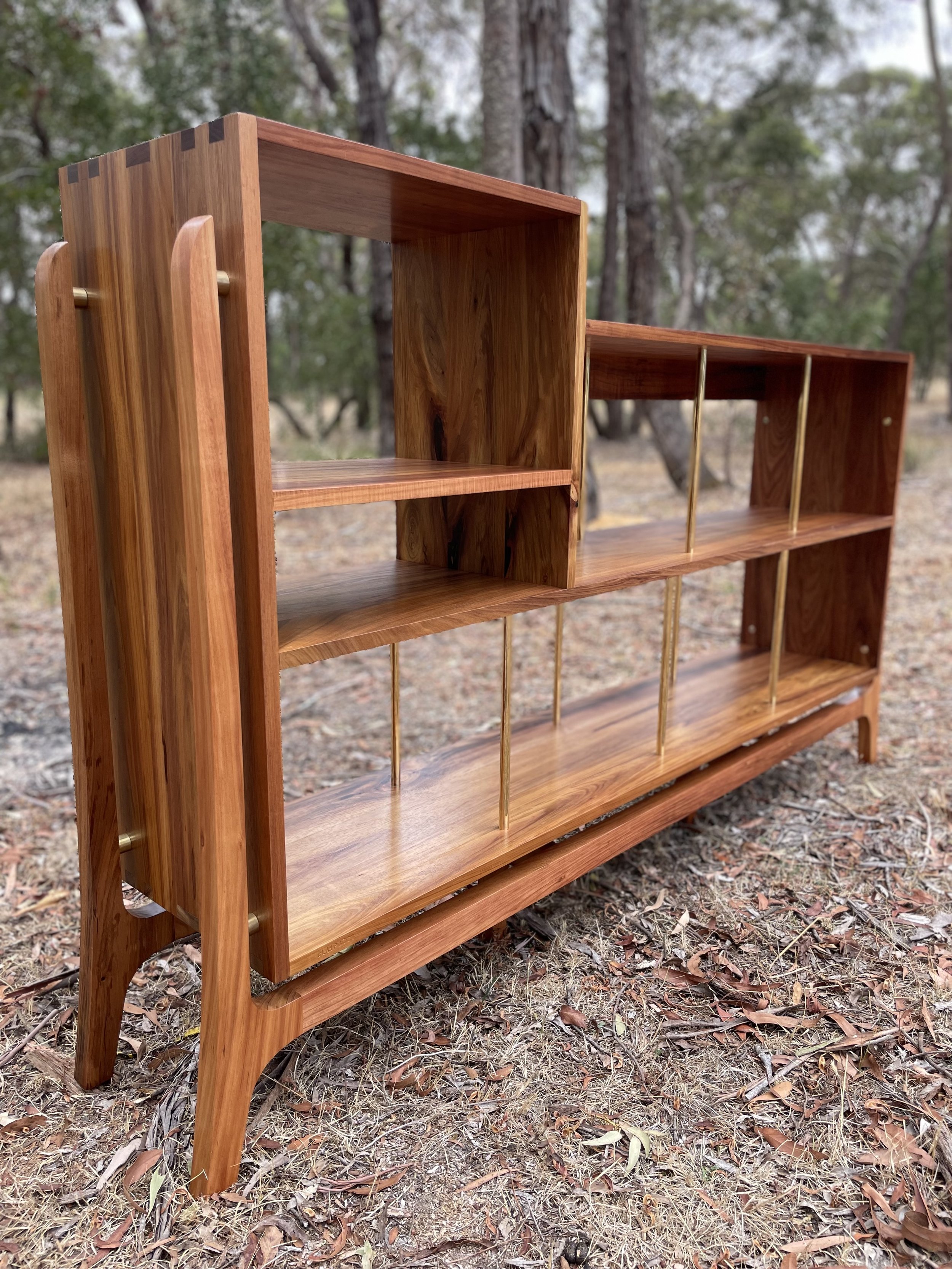 A wooden bookshelf with multiple shelves and gold-colored support rods is placed outdoors on dirt and fallen leaves, with trees in the background.