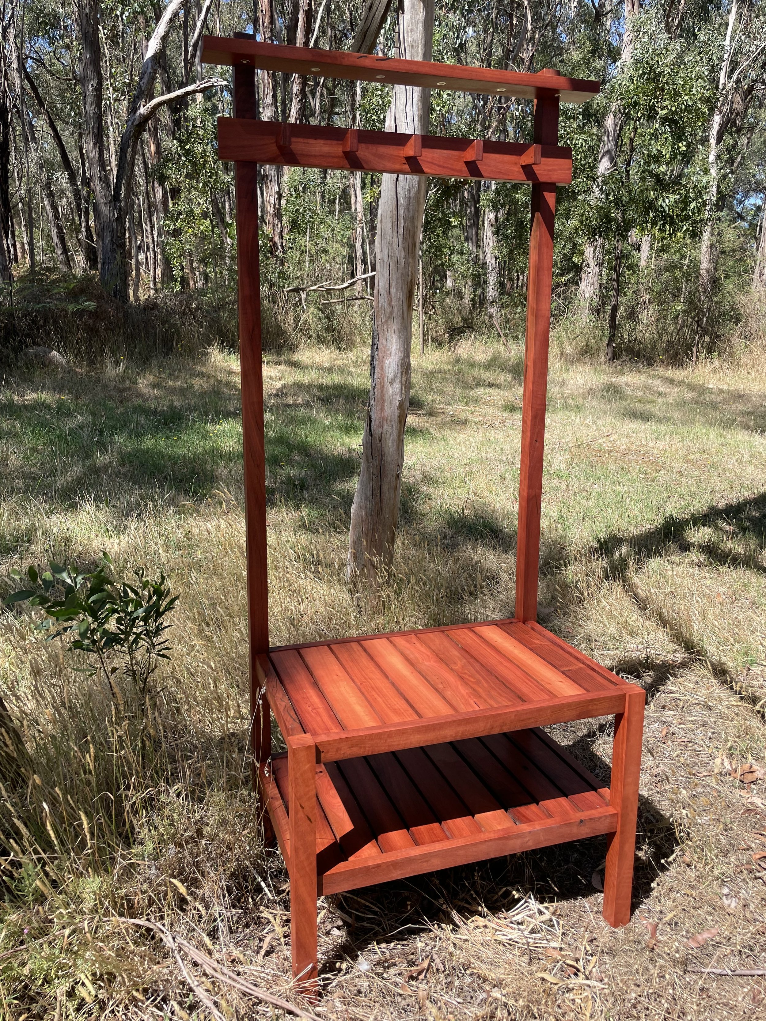 A wooden outdoor structure with a square platform at the bottom and a horizontal bar at the top, located in a forested area with trees and grass.