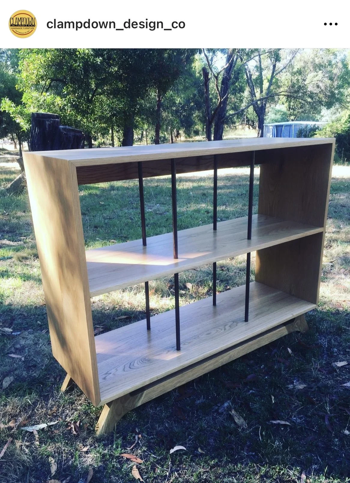 A wooden bookshelf outdoors on grass with trees in the background, featuring a modern design with metal supports.
