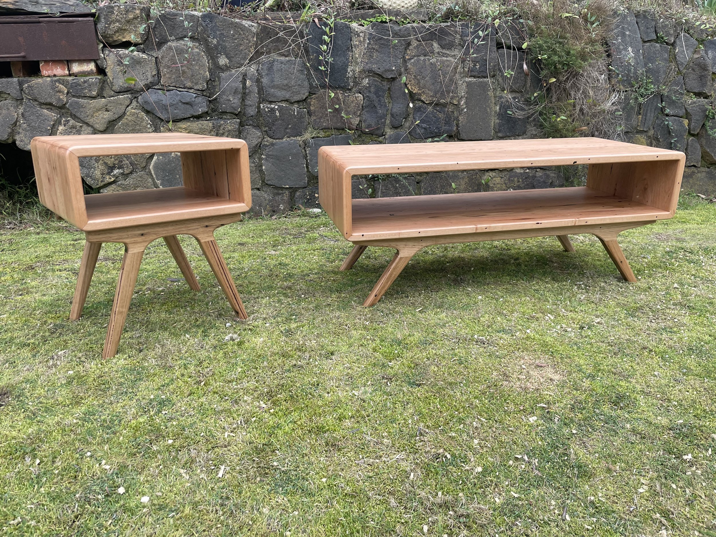 Two wooden furniture pieces, a small square side table and a larger rectangular coffee table, are placed on a grassy area in front of a stone wall. Both have a minimalist design with angled legs.