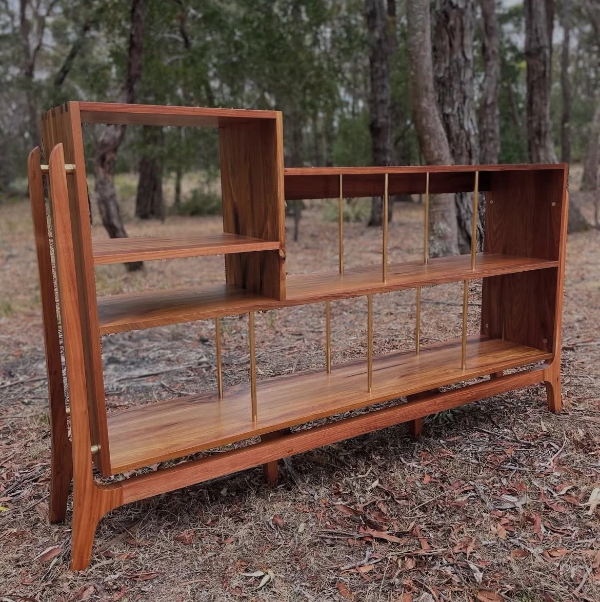 A wooden bookshelf with multiple shelves and vertical dividers, standing outdoors on a dirt ground with trees in the background.