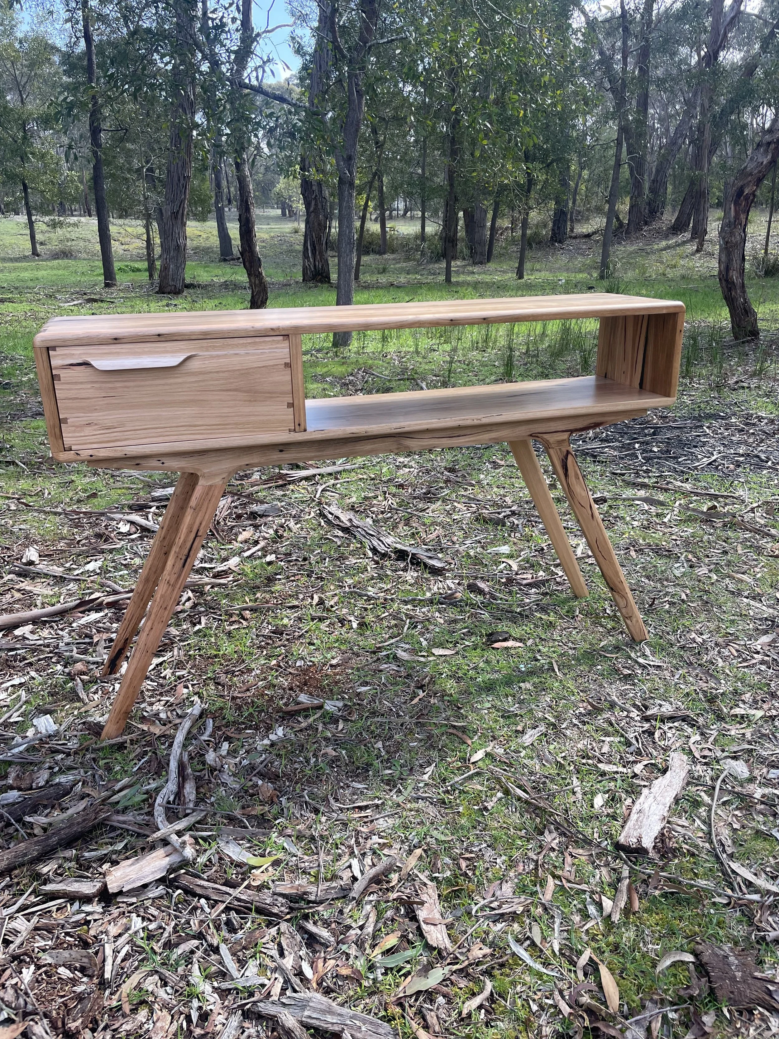 A timber hall stand with one drawer and an open shelf, set outdoors in a wooded area with green grass and trees.