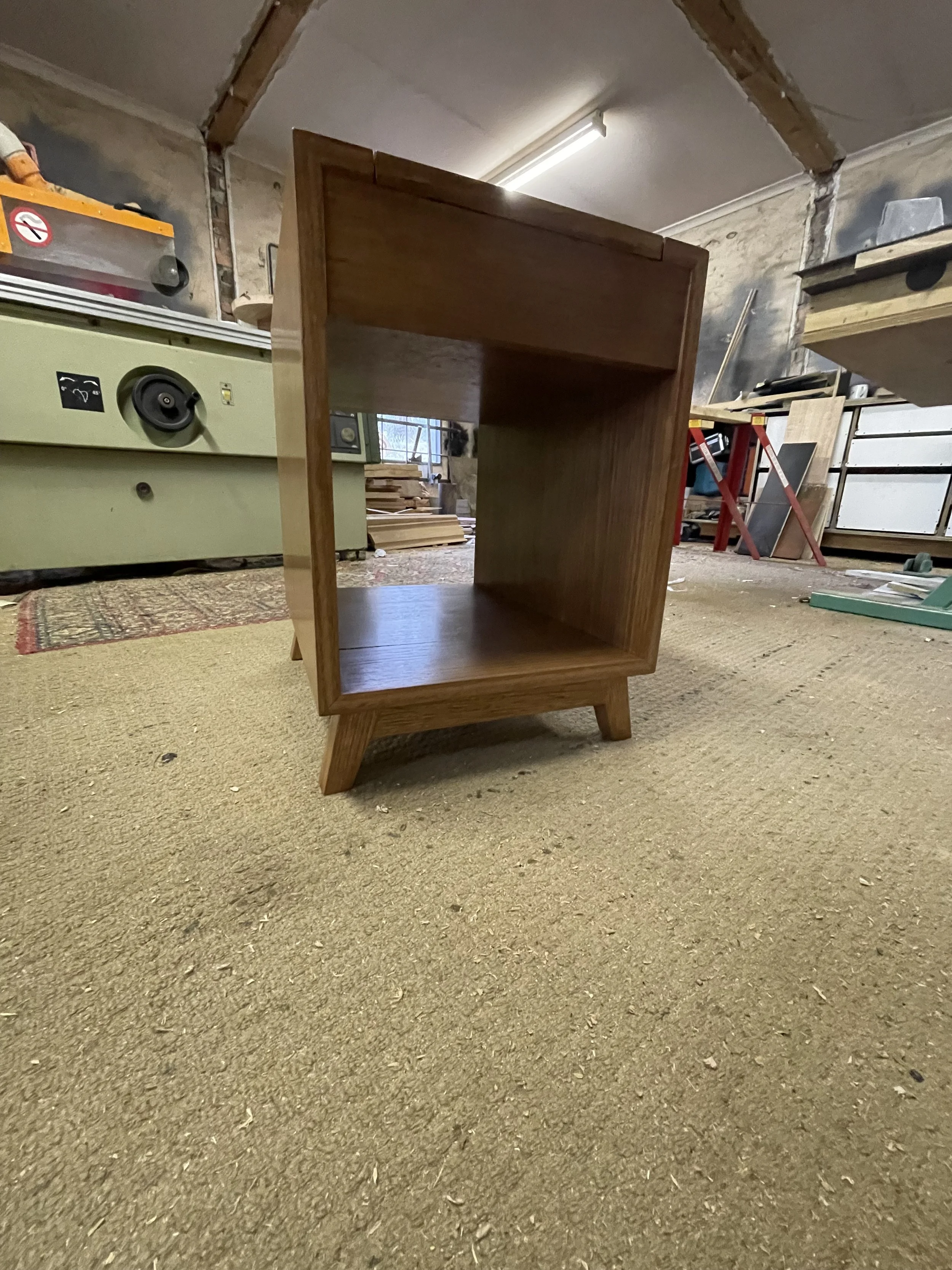 A wooden bookshelf with an open front, standing on angled legs in a woodworking workshop.