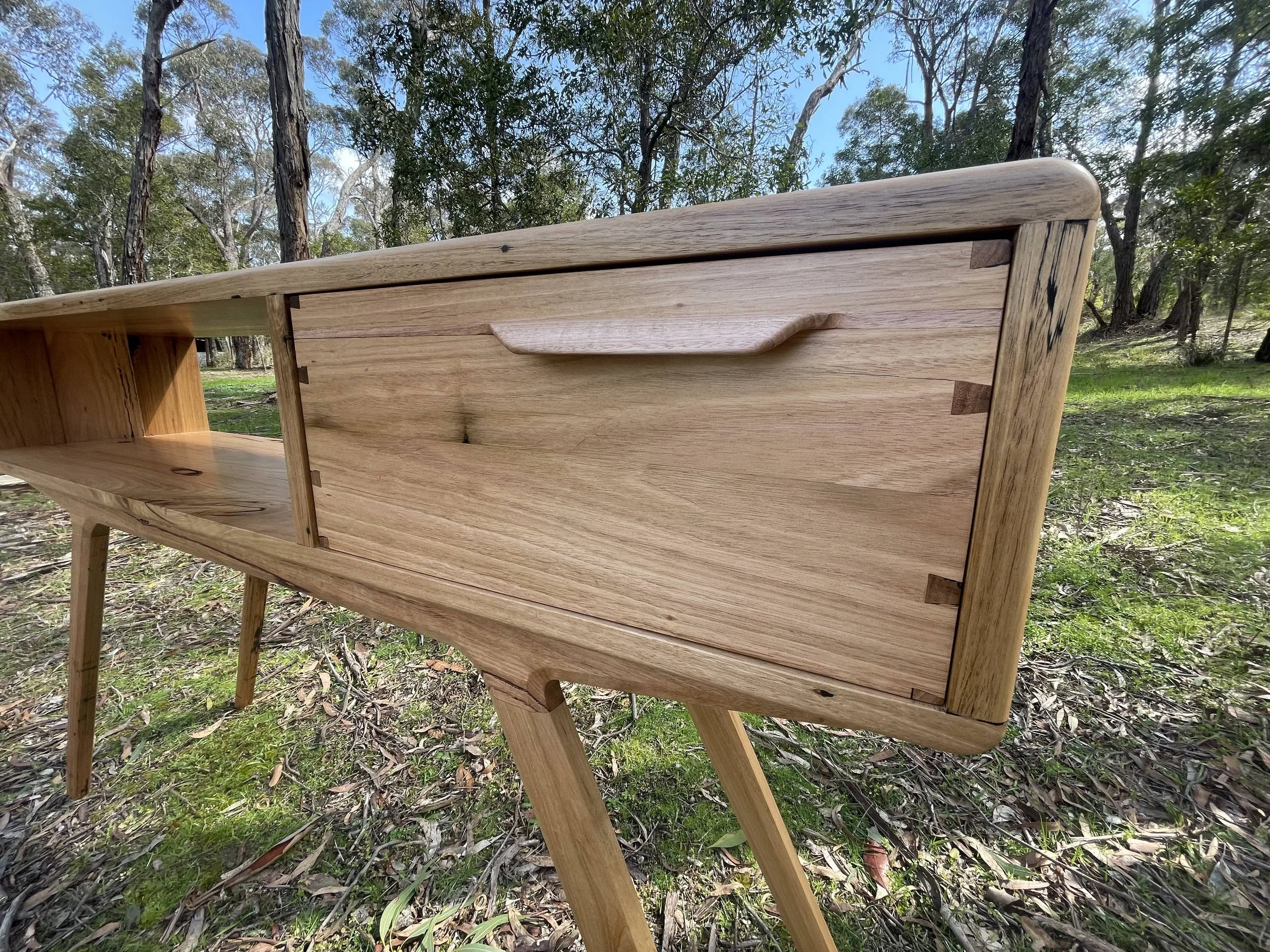 A wooden piece of furniture, possibly a cabinet or sideboard, with a drawer and open shelves, set outdoors in a natural wooded area.