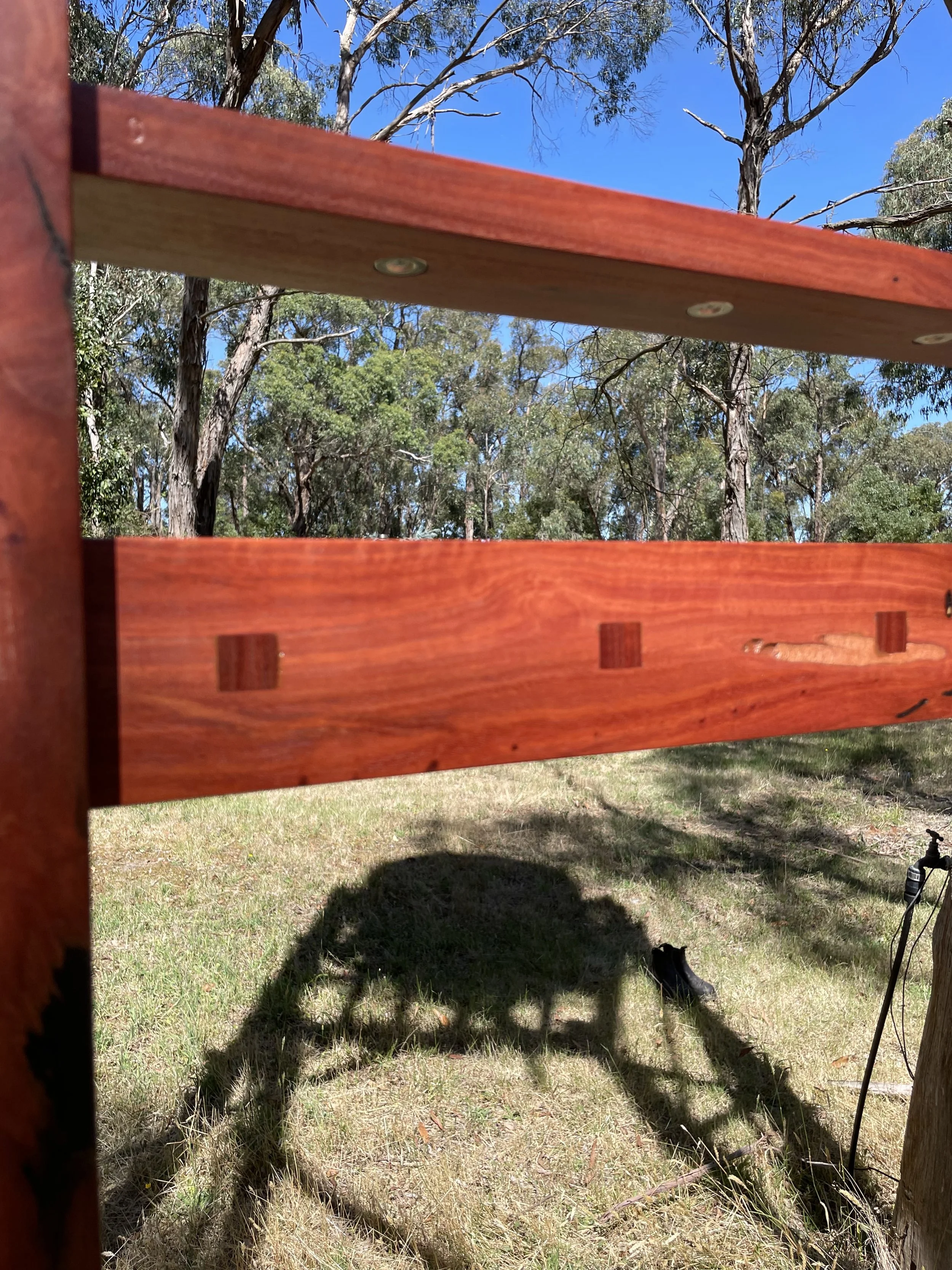 Close-up of a wooden structure outdoors with trees and blue sky in the background, casting a shadow on the grass.