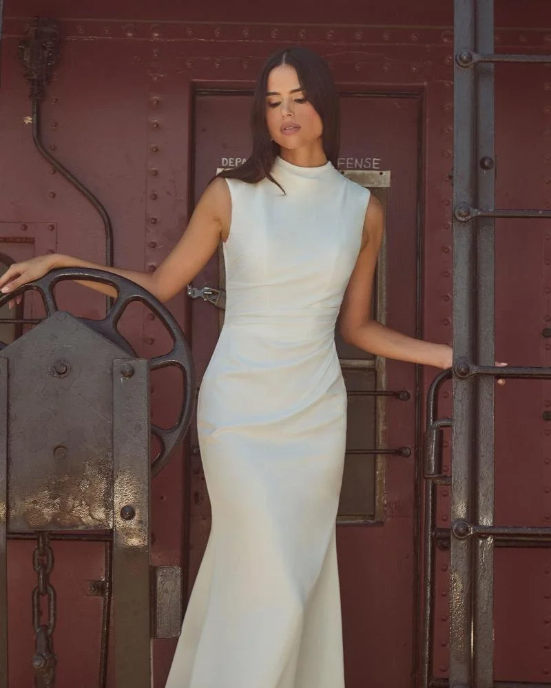 La Perle Maeve modern minimalist satin sheath wedding dress with high mock neckline, cap sleeves, and stunning open back with button details.