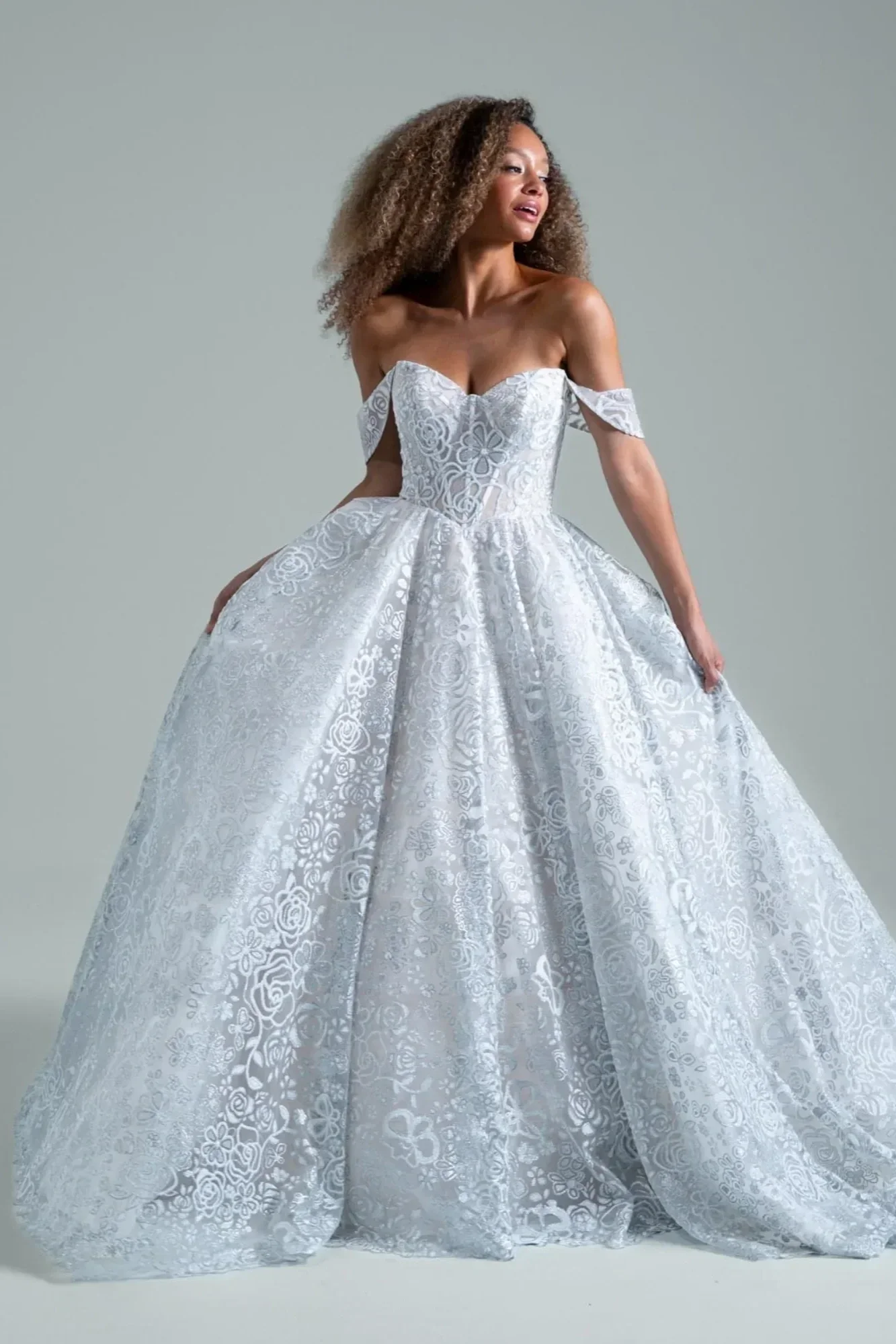 Woman wearing a white off-shoulder, lace wedding gown with a full skirt, standing against a plain background.