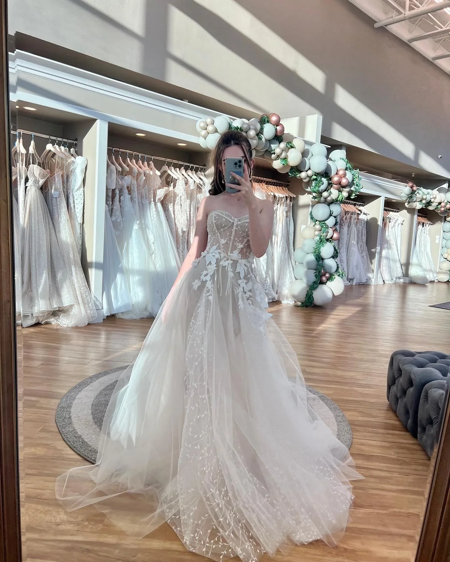 A woman trying on a strapless wedding dress with floral embroidery in a bridal boutique, taking a selfie in a mirror. The boutique has wedding dresses on racks, decorative balloon arrangements, and a wooden floor.