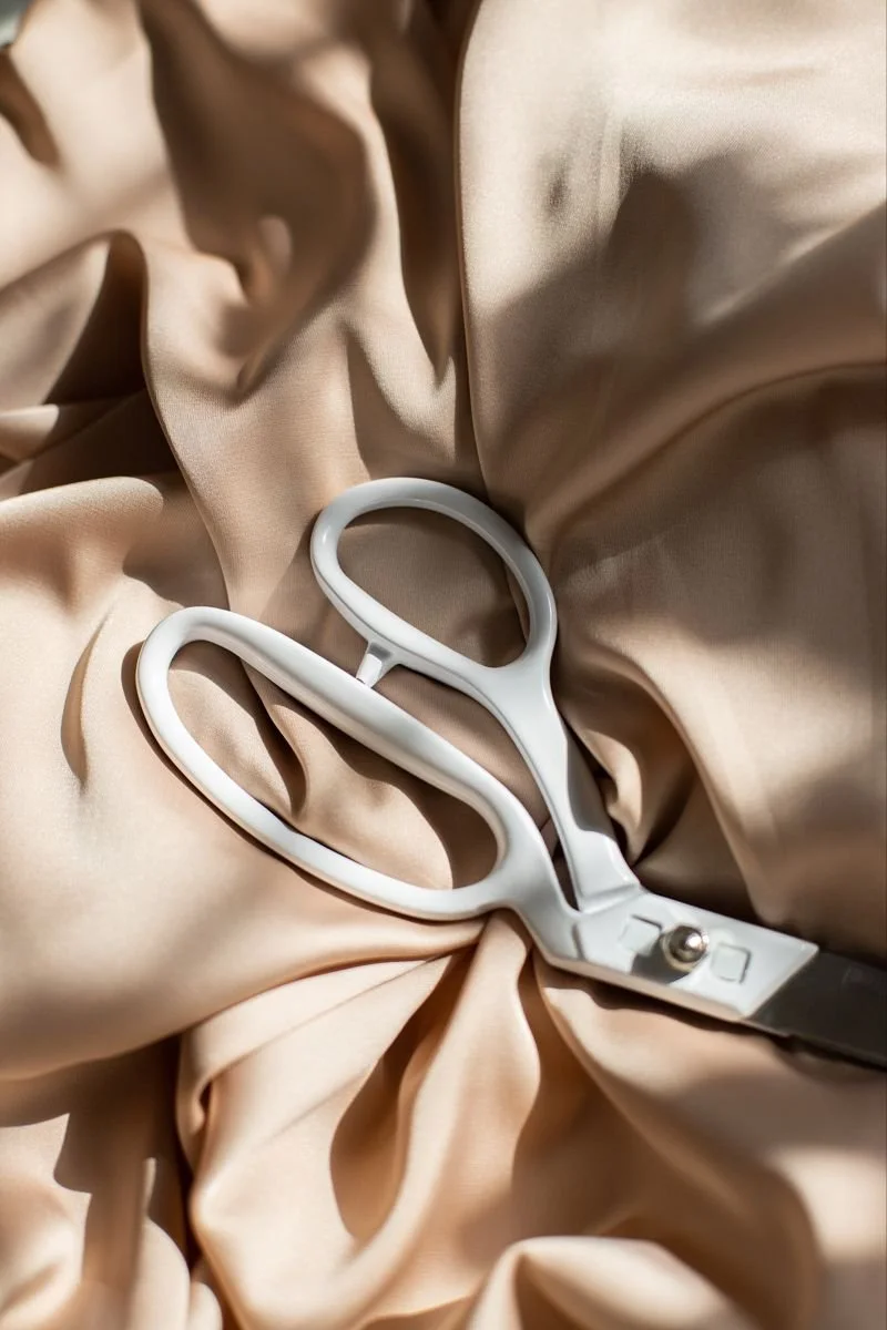 A pair of white scissors resting on beige fabric.