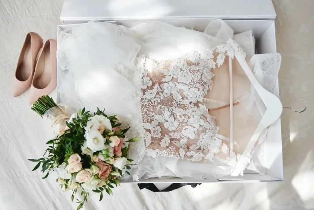 A wedding dress with lace detail in a box, a bouquet of white and pink roses, and a pair of nude-colored high heels.