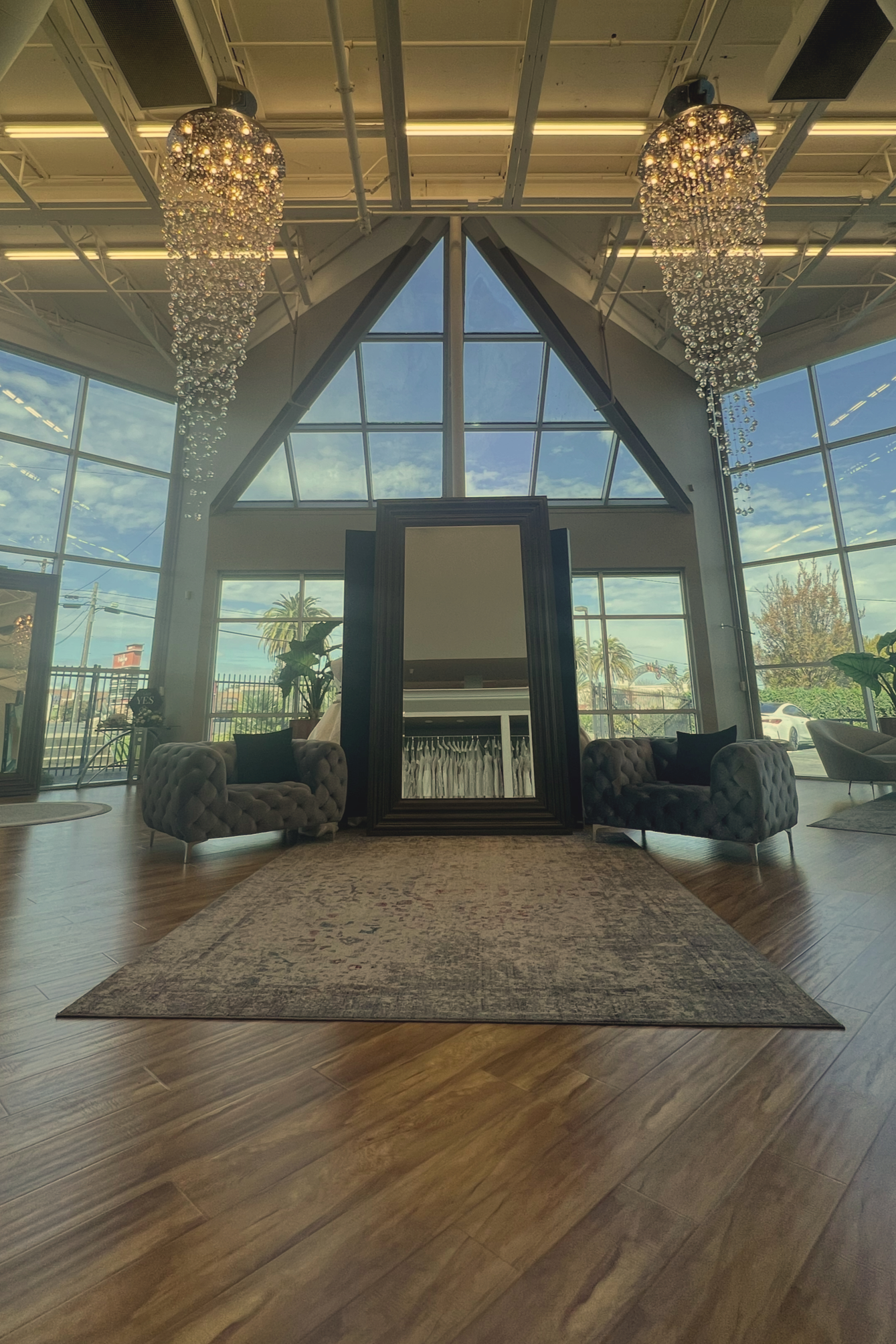 Interior of a modern lounge or waiting area with large windows, a scenic view, two tufted armchairs, a mirror, chandeliers, and a rug on a wooden floor.