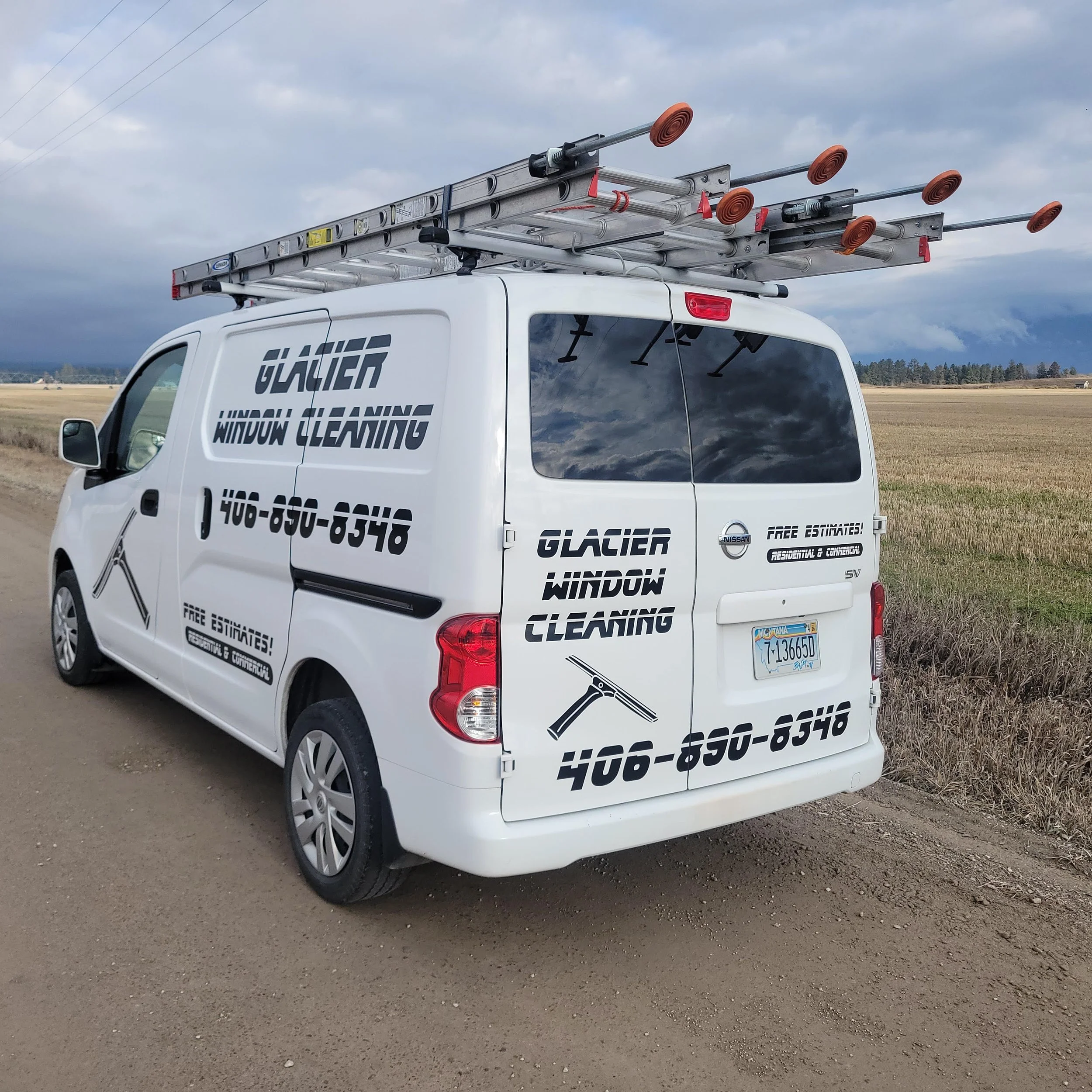 White van with 'Glacier Window Cleaning' and phone number on the side and back, ladder on the roof, parked on a dirt road in a rural area with fields and cloudy sky in the background.