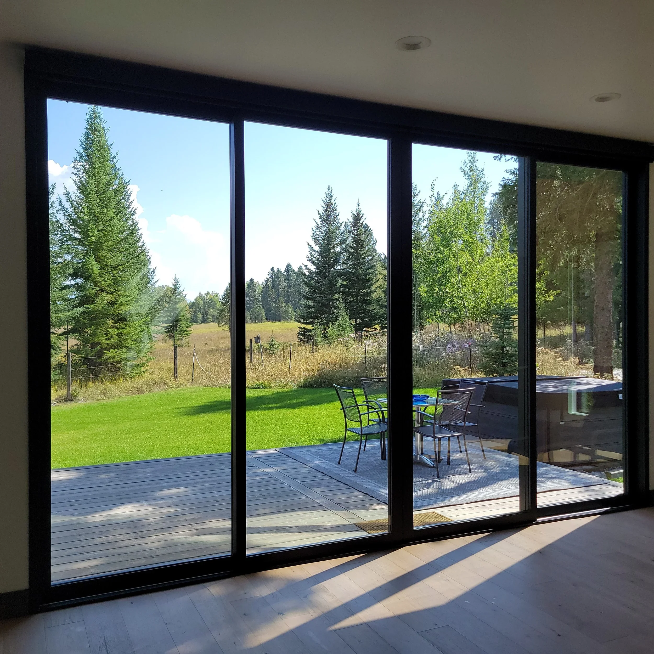 View through large sliding glass doors showing a backyard with a wooden deck, patio furniture, a hot tub, green grass, and tall trees under a blue sky.