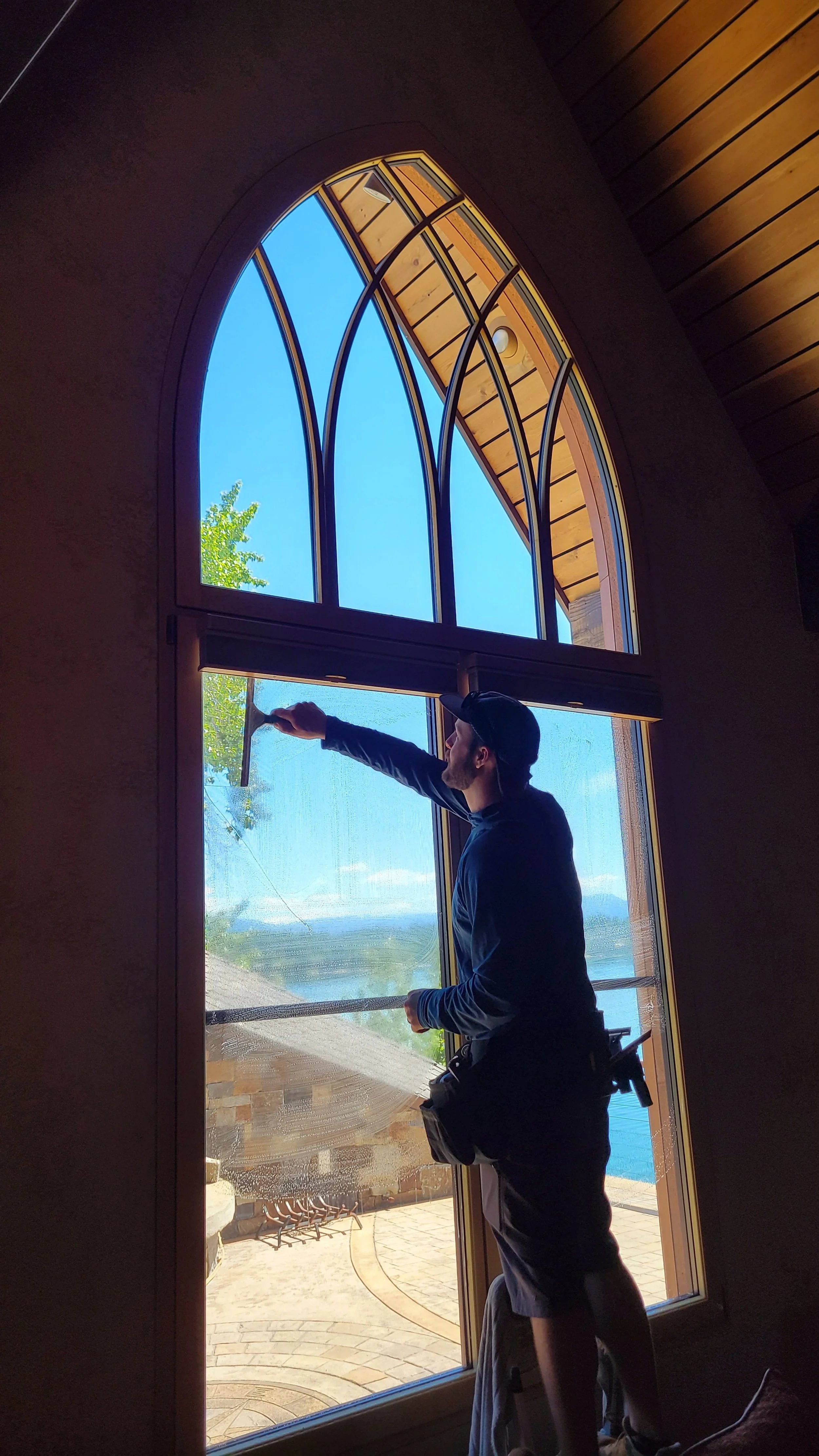 A man cleaning large arched window inside a house with a squeegee, with a view of trees and water outside.