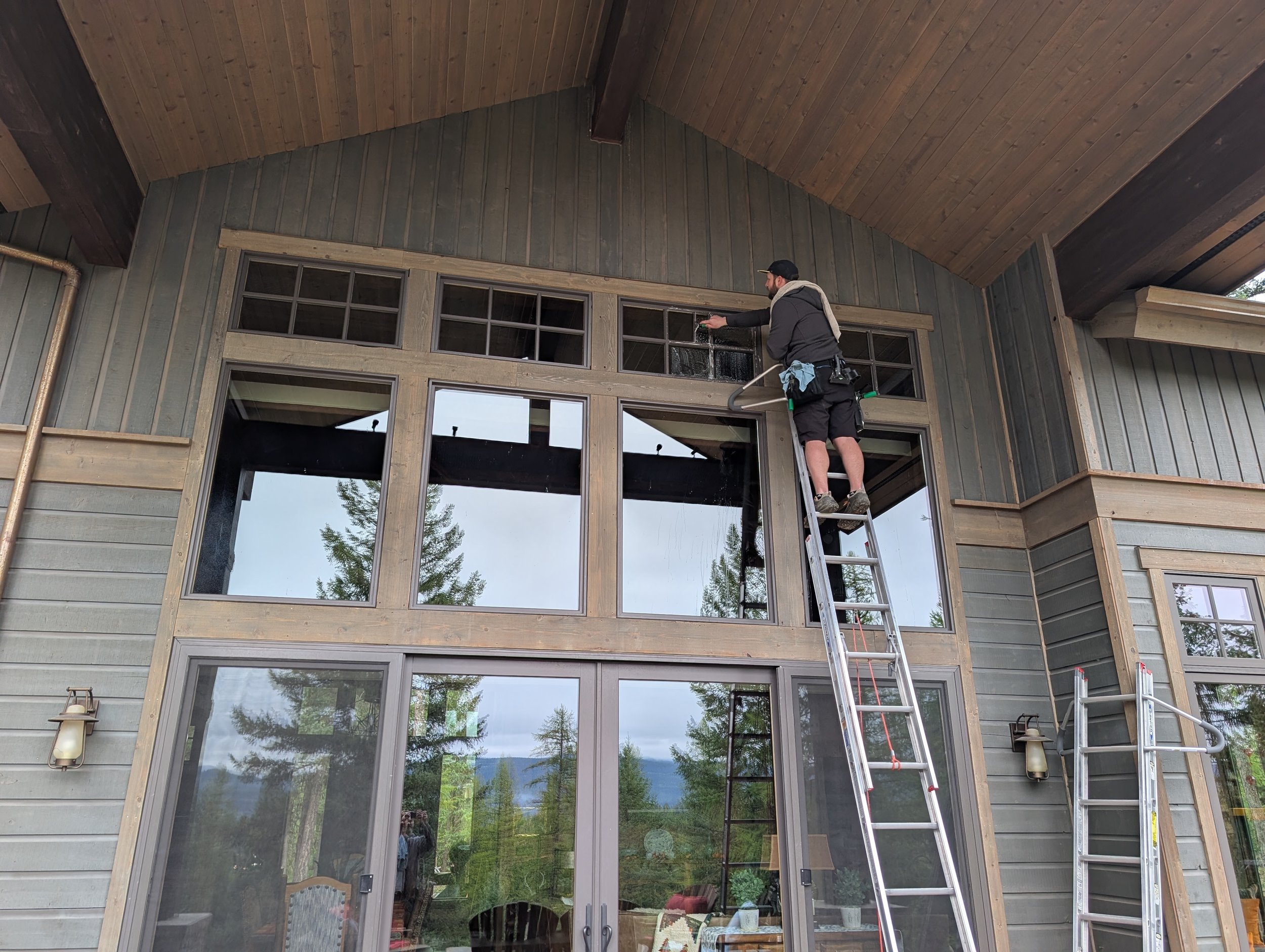 A man on a ladder cleaning windows on a large, multi-pane glass door and window section of a house with wooden and gray siding.