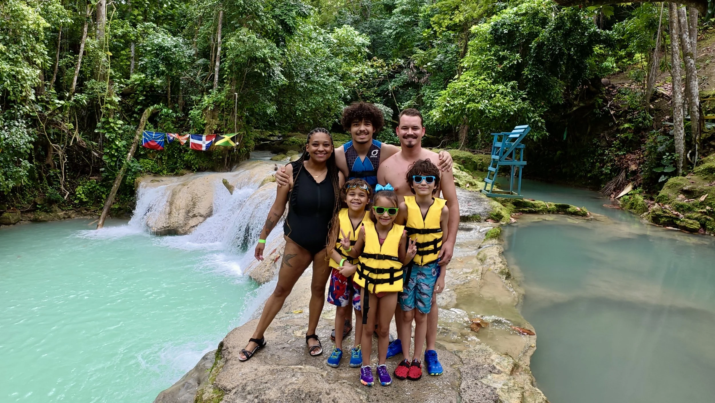 Group of seven people, including children and adults, wearing life jackets and swimming gear, standing on rocks near a waterfall and river in a lush green jungle setting, with flags hanging in the background.