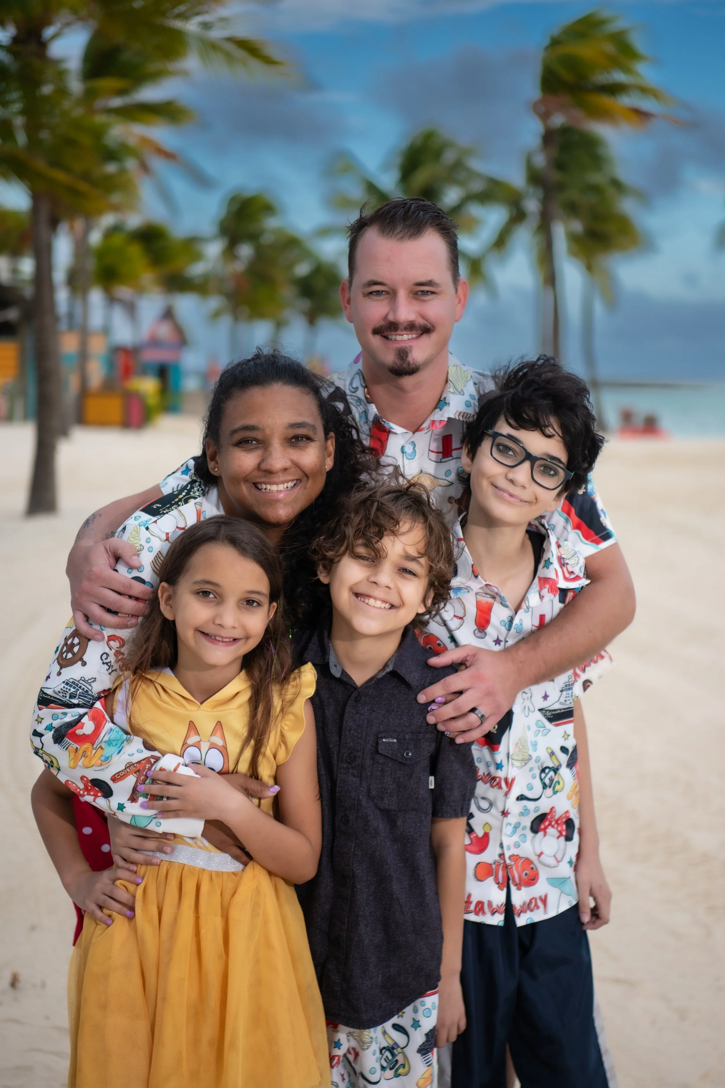 A happy multiracial family of six enjoying a day at the beach, standing close together with a backdrop of palm trees and blue sky.