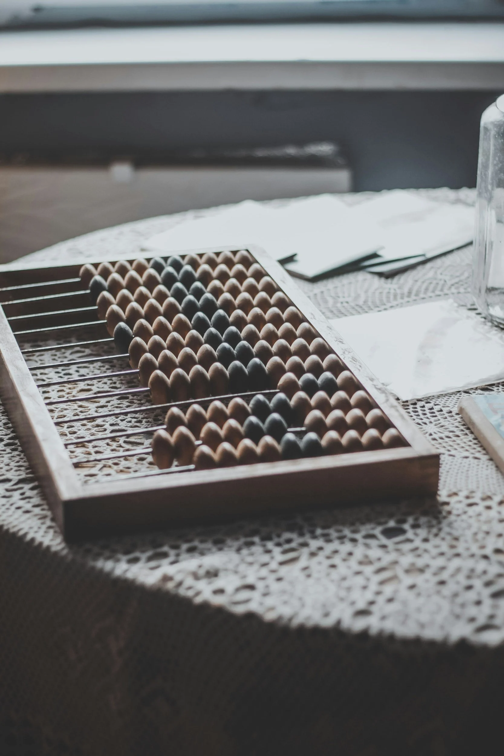 A wooden tray containing brown and black eggs placed on a table with a lace tablecloth, with some papers and a glass container nearby.