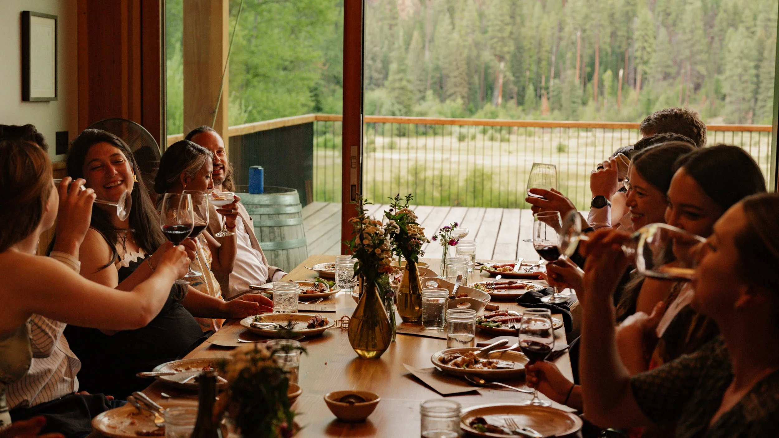 Group of people enjoying a meal together at a long dining table in a room with large glass windows overlooking a forested landscape. Minam Lodge Wallowas