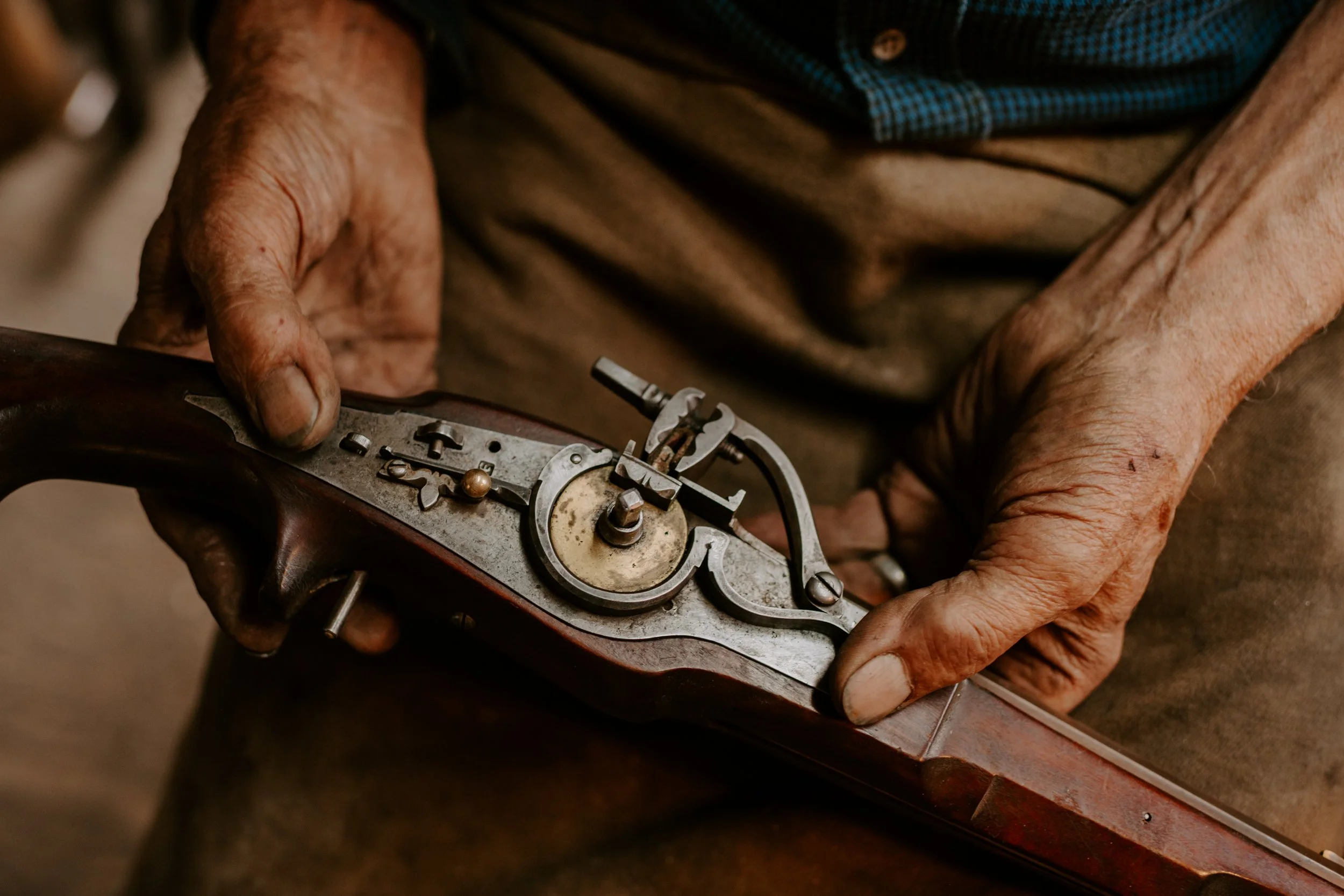 Close-up of elderly person's hands working on the mechanical parts of a vintage gun or object, showing detailed metal components and aged skin.