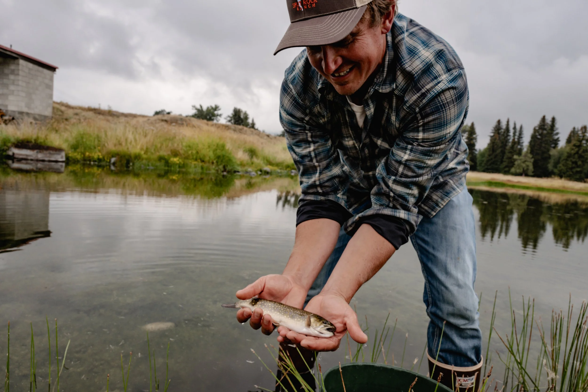 A man in a plaid shirt and baseball cap holding a freshly caught fish near a body of water in a natural outdoor setting with trees and a cloudy sky in the background. Wallowa River Joseph Oregon Nez Perce Tribes.