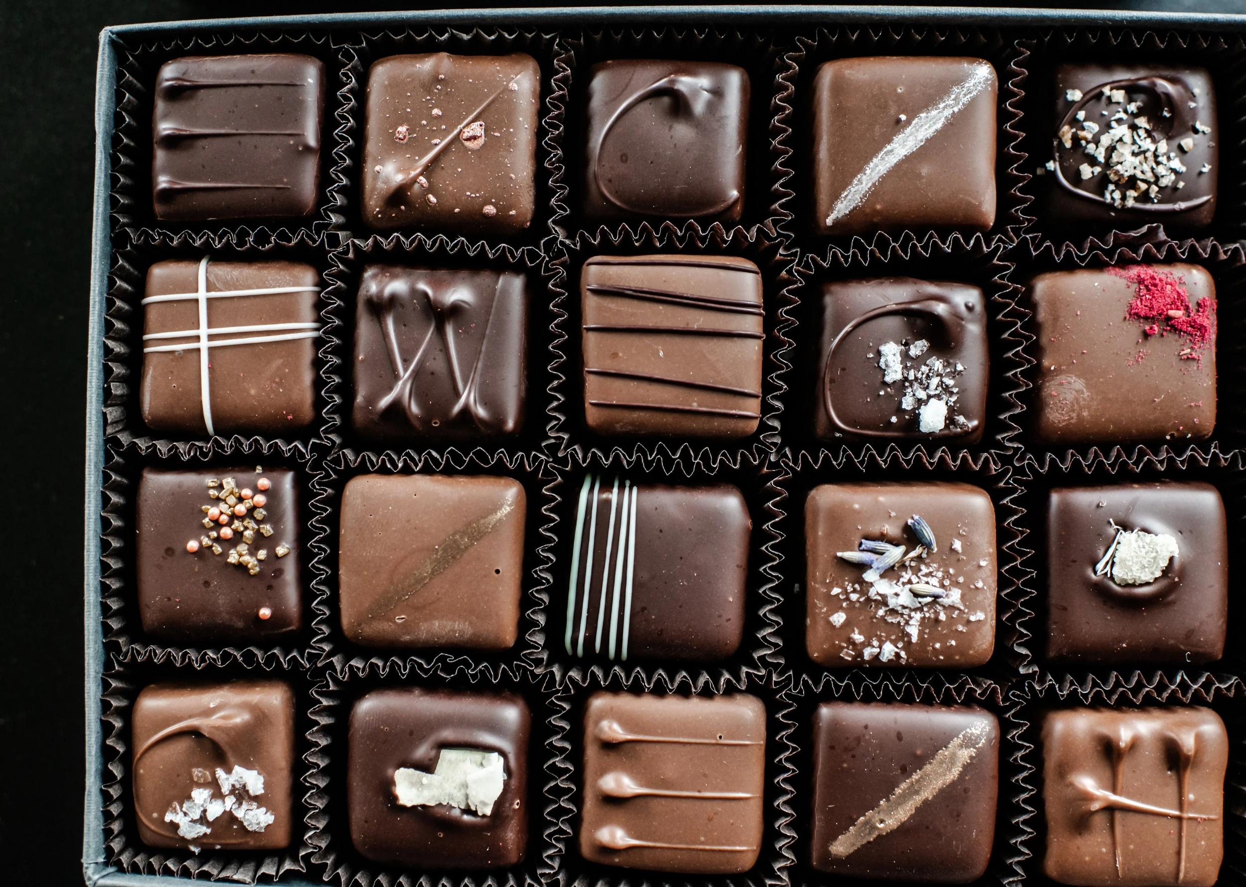 A box of assorted milk chocolates topped with various decorations like sprinkles, drizzles, and sea salt, arranged in individual compartments.