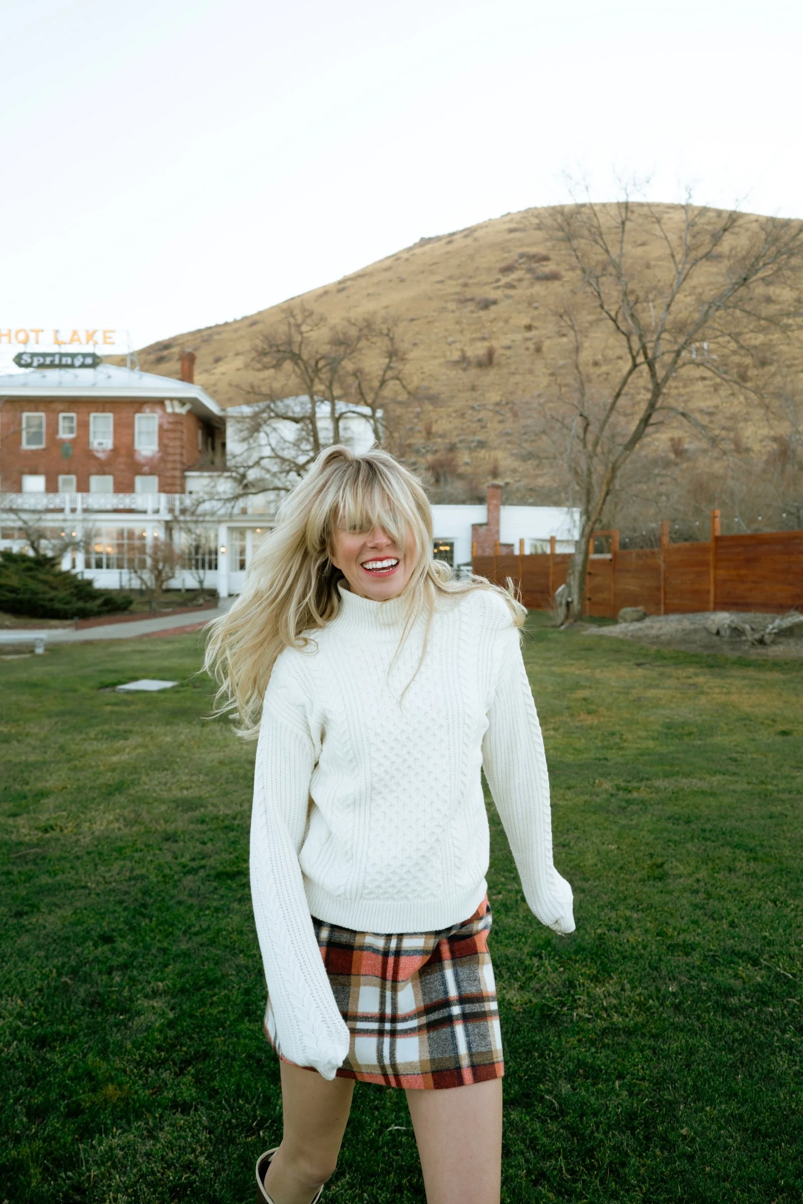 A young woman with long blonde hair smiling, wearing a cream-colored turtleneck sweater, plaid shorts, and black shoes, standing outdoors on a grassy area with a hill, leafless trees, and a building with a sign that reads "HOT LAKE" and "Spring" in the background.