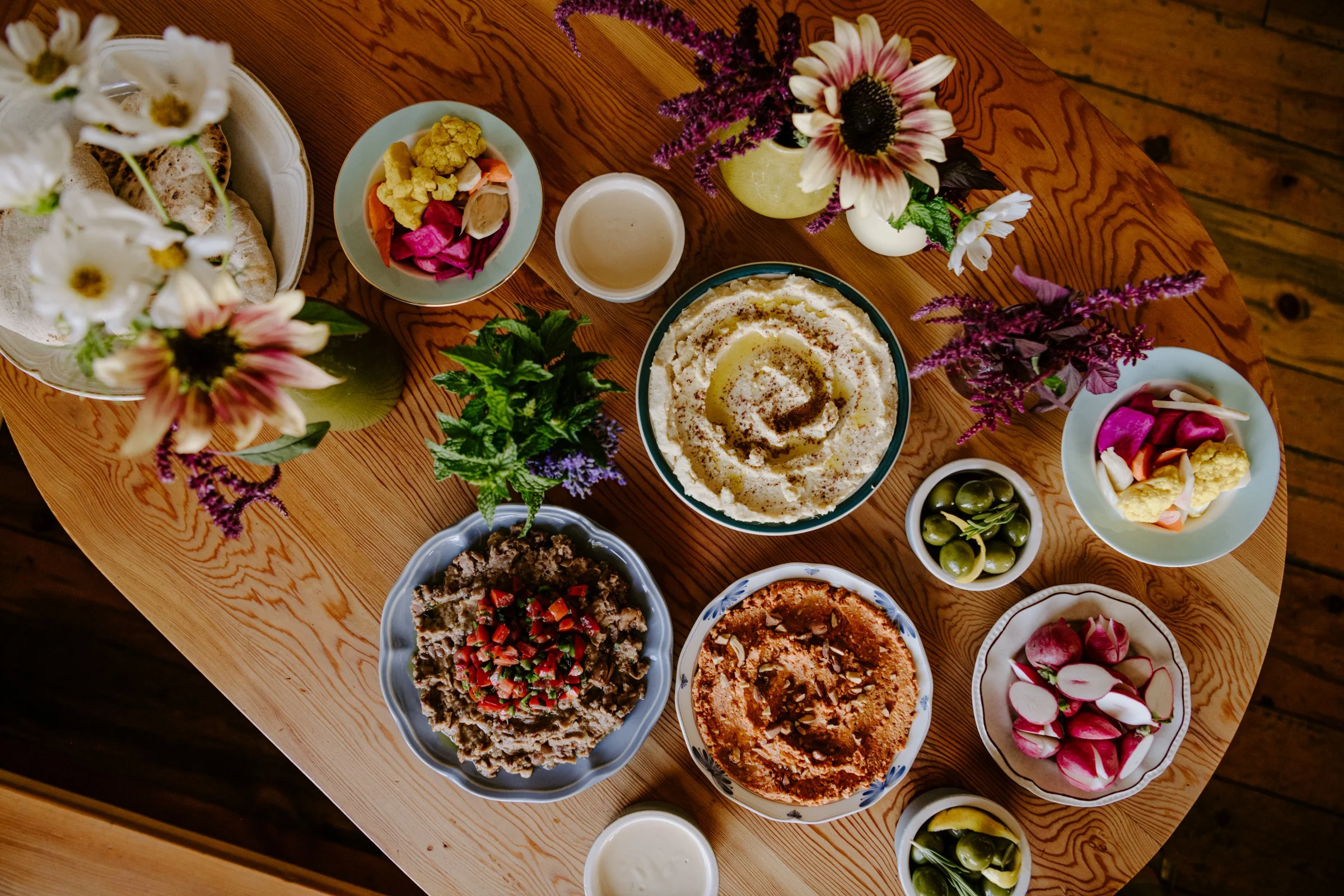 A wooden table set with various dishes including dips, salads, a floral centerpiece, and small bowls of olives and radishes, surrounded by colorful flowers in vases.
