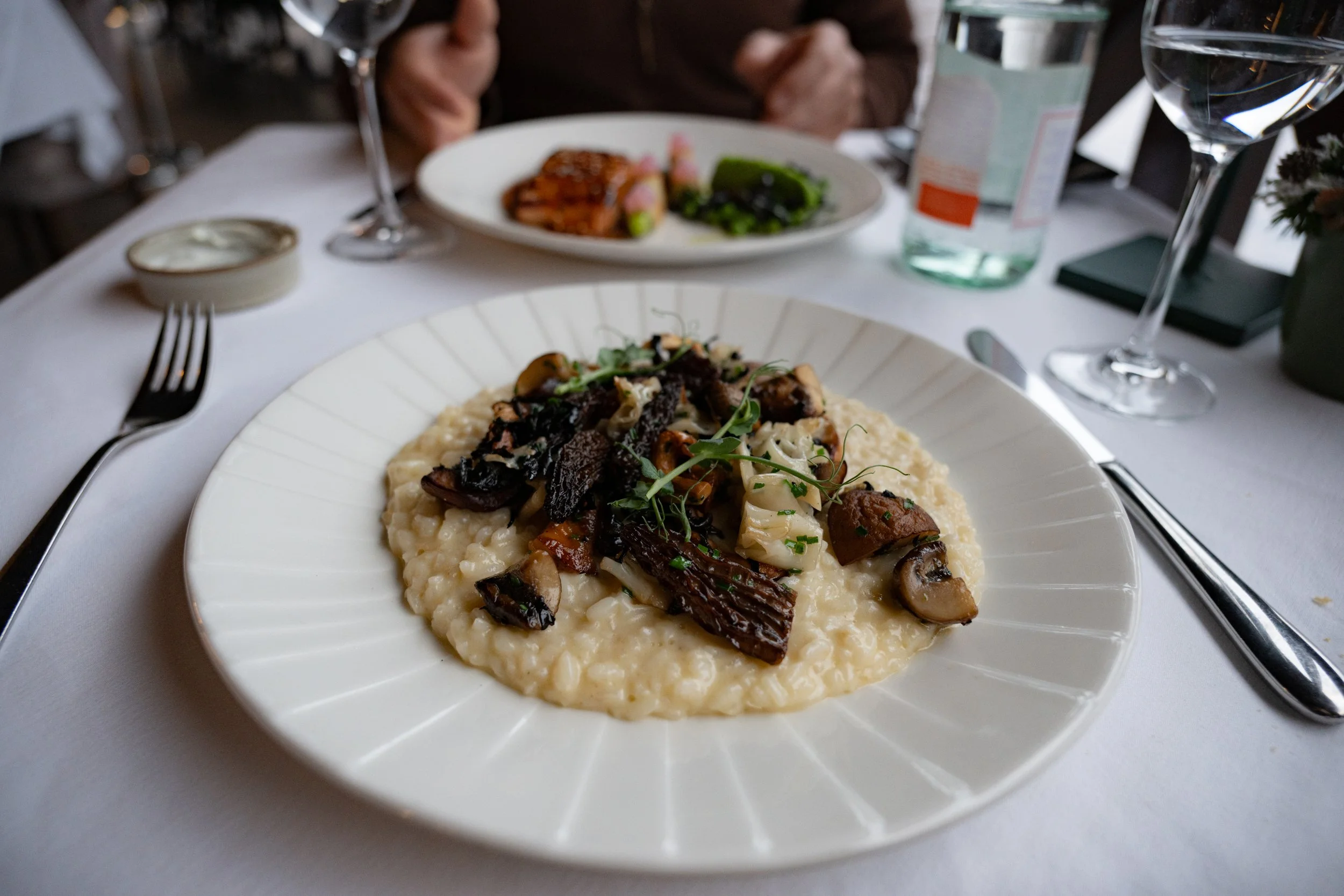 A white plate of risotto topped with sautéed mushrooms, herbs, and dark vegetable bits, on a white tablecloth, with wine glasses, a fork, and a small dish nearby, at a dining setting with people in the background.