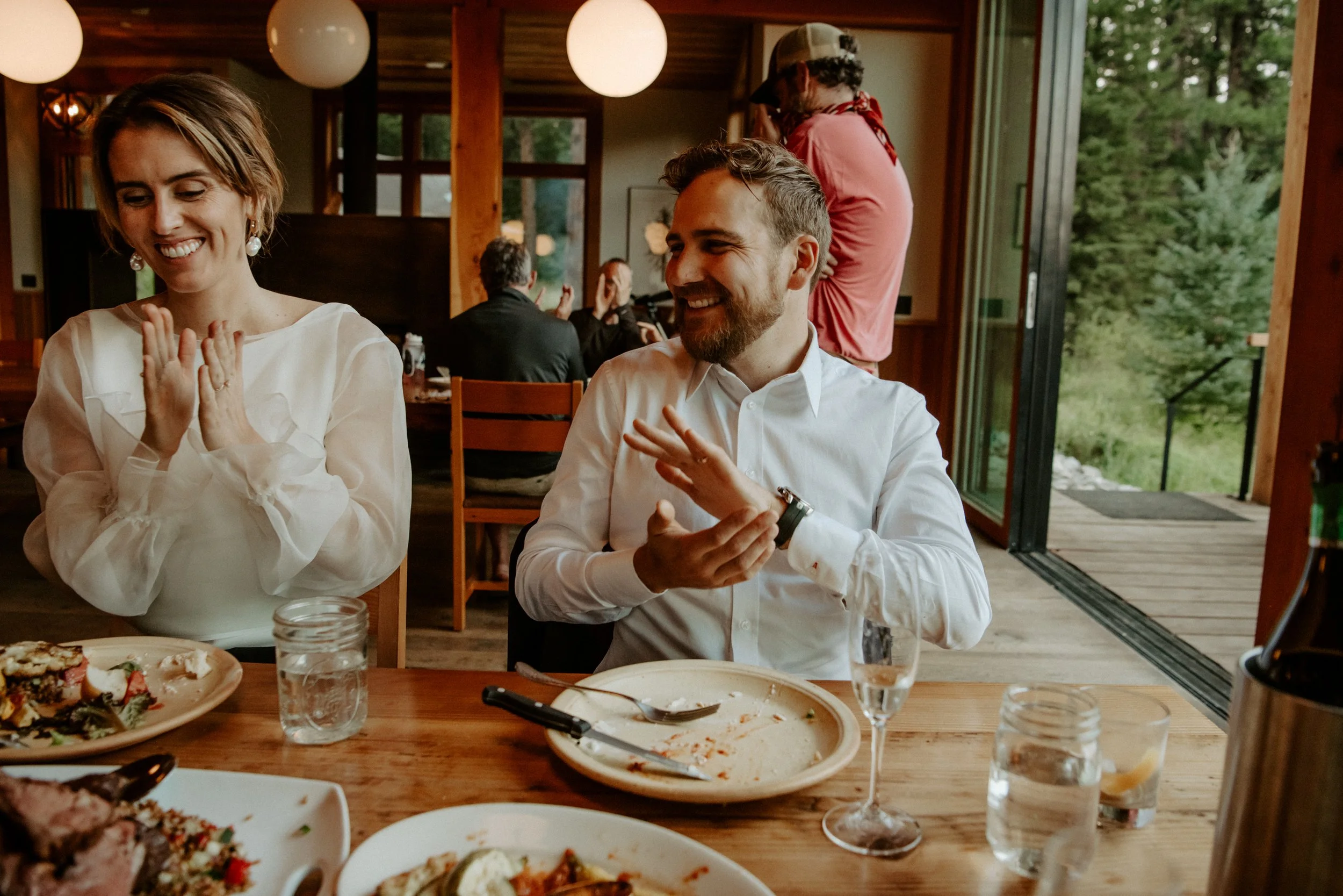 People enjoying a meal at a restaurant or cafe with a wooden interior and large windows, surrounded by greenery outside.