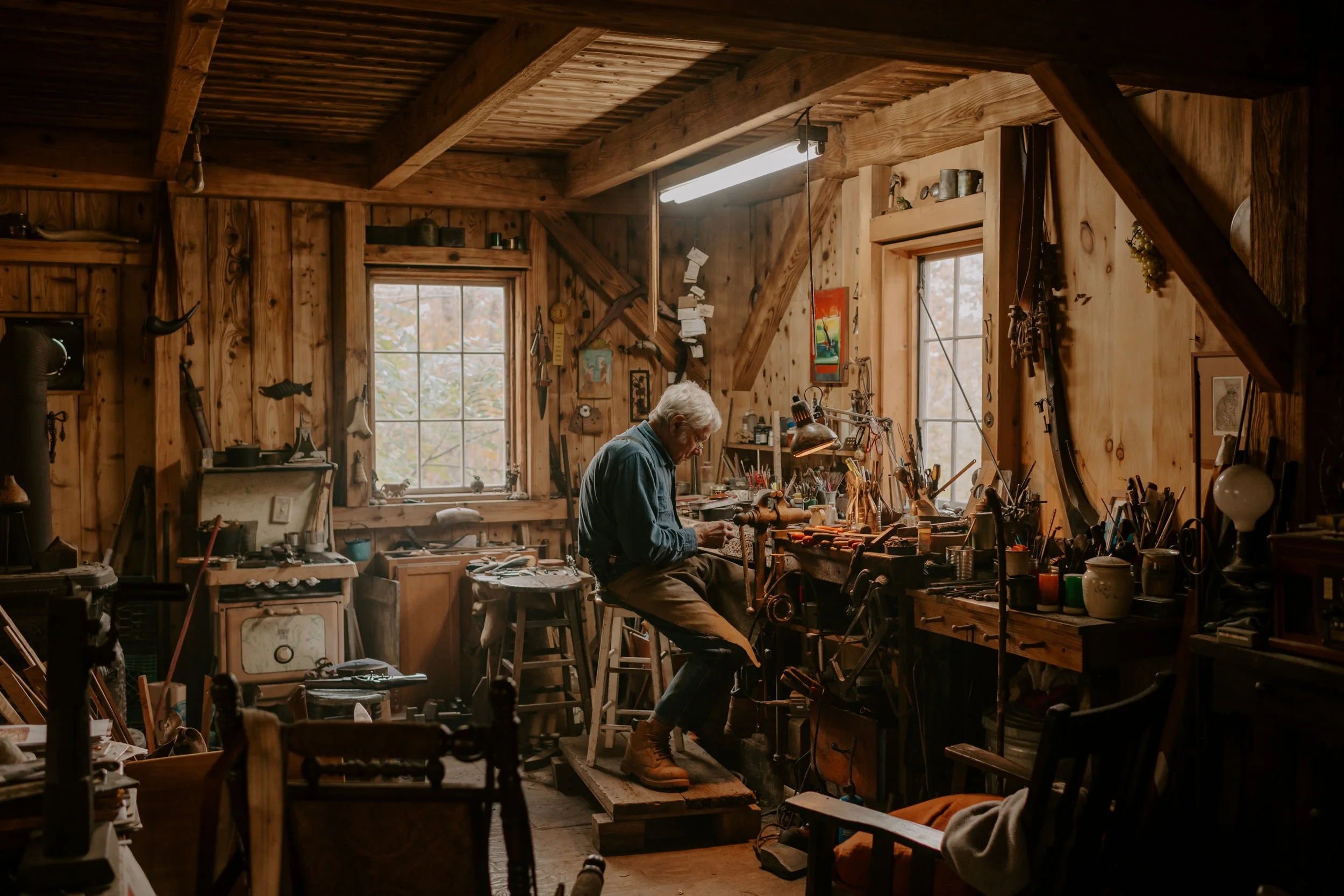 An elderly man working in a rustic woodworking workshop filled with tools and wooden furniture.