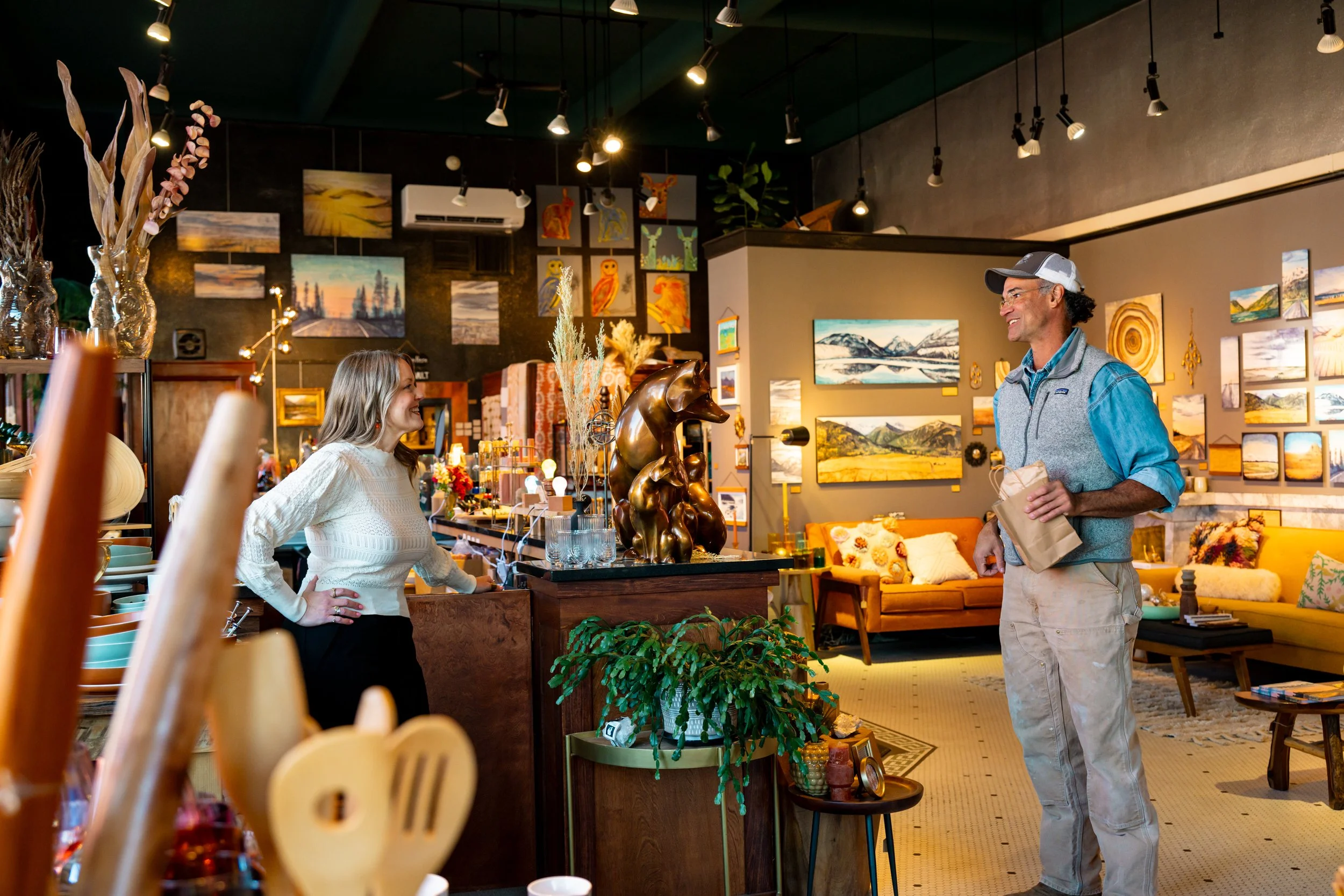 A woman and a man are conversing inside an art gallery or home decor store. The woman is wearing a white sweater and has her hand on her hip, smiling at the man. The man is wearing a gray vest, blue shirt, and a baseball cap, holding a paper bag and 