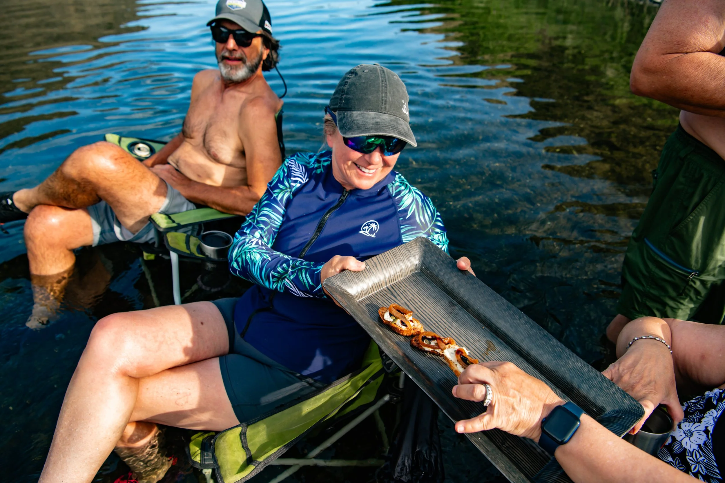 Smiling woman in blue jacket and sunglasses sitting in a kayak, holding a tray of grilled sliders, with a man sitting behind her in a life jacket, on a calm lake with trees in the background.