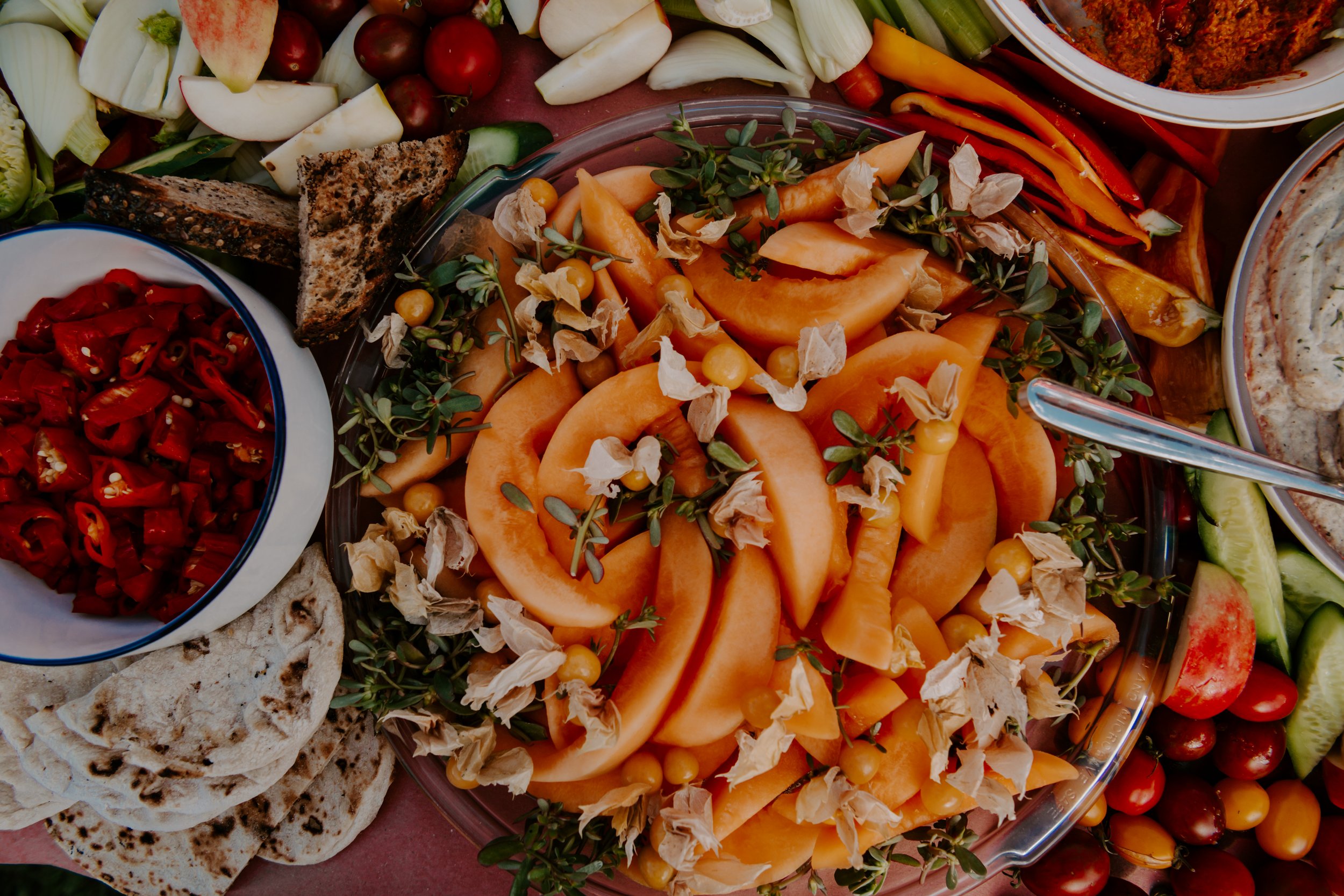 Assorted fresh fruits and vegetables on a table, including sliced melon, cherry tomatoes, cucumbers, red and yellow peppers, and greens, along with bread and dips.