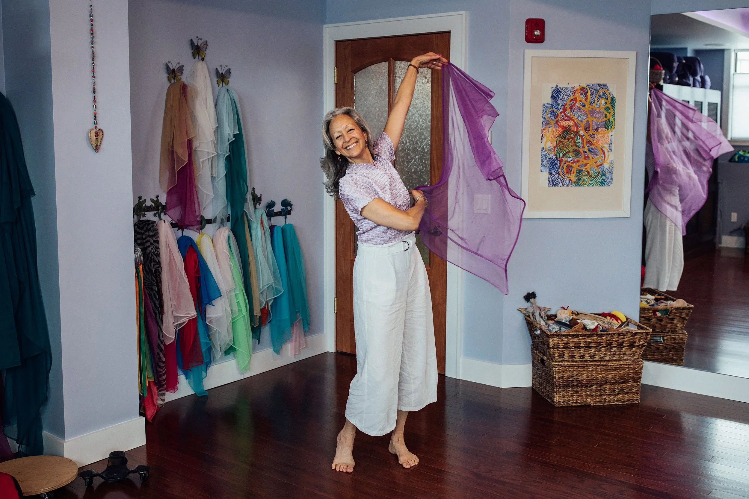 A smiling woman standing in a room with colorful dresses and tulle skirts, holding purple fabric. The room has hardwood floors, wall art, baskets of sewing or craft supplies, and a mirror.