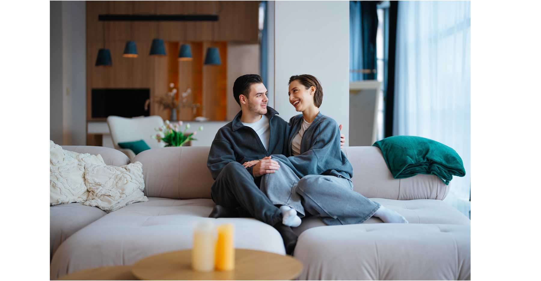 A young couple sits on a light-colored sofa in a modern living room, smiling and looking at each other. The man has dark hair and is wearing a navy jacket over a white shirt, and the woman has short brown hair and is wearing a gray hoodie. There are candles and pillows on the sofa, a folded blanket on the right armrest, and a window with sheer curtains letting in natural light.