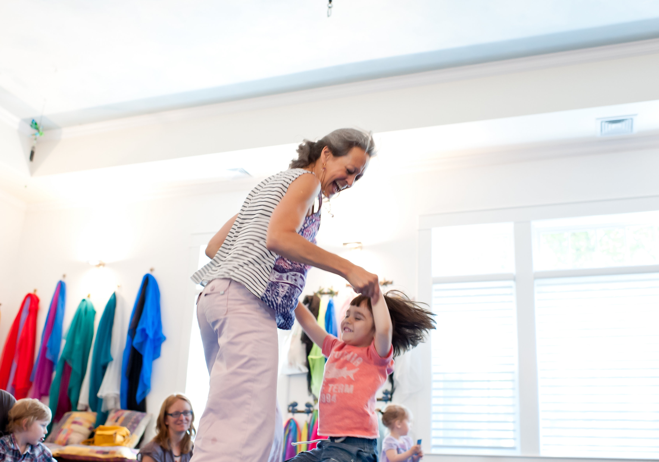 Woman and girl dancing and playing together indoors, with other children and adults in the background.
