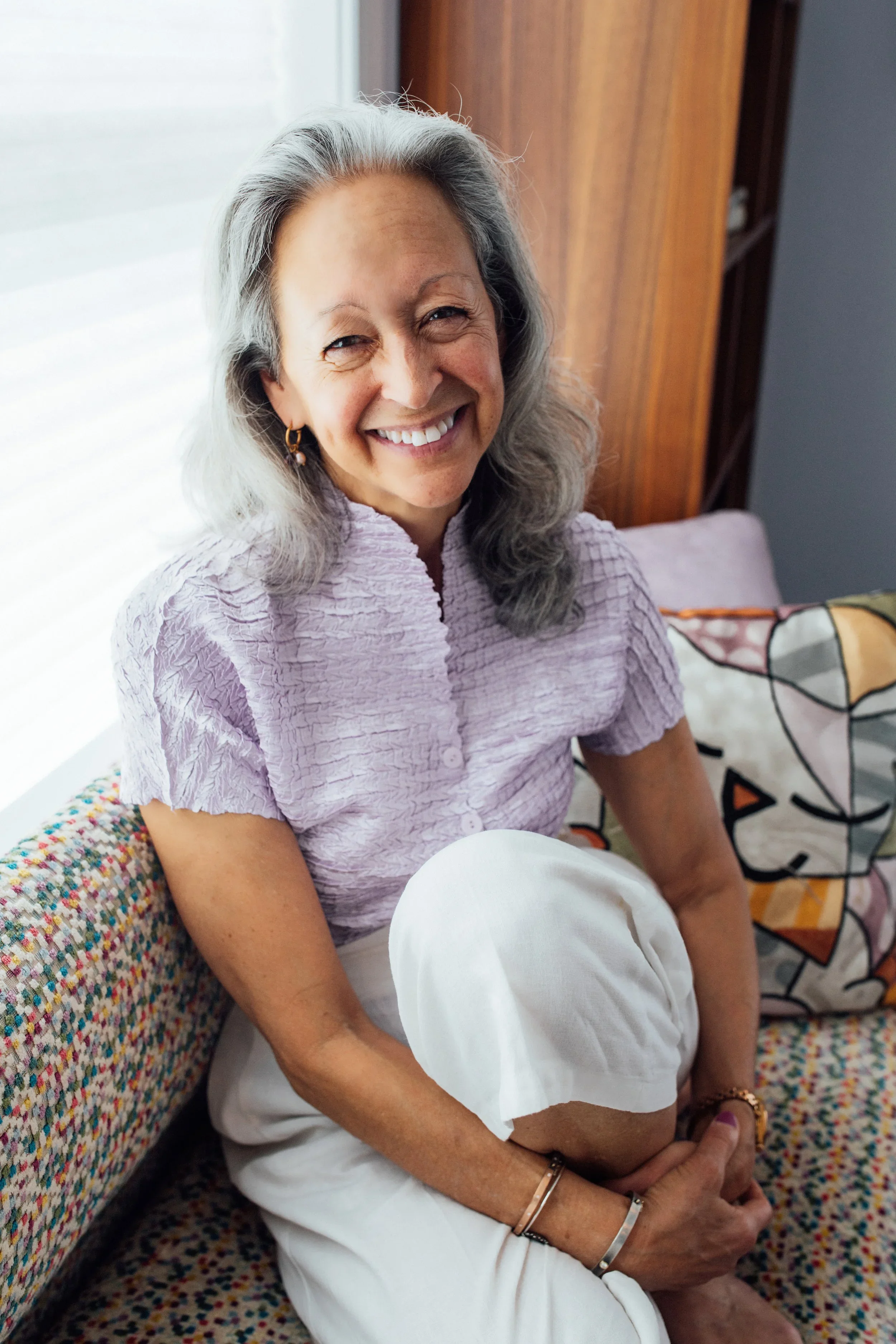 Senior woman with gray hair smiling while sitting on a couch with a colorful cushion, holding her knee, in a well-lit room.
