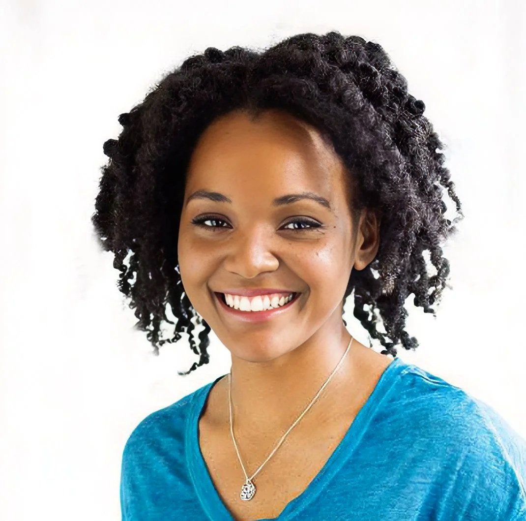 A woman with curly black hair smiling, wearing a blue shirt and a silver necklace, against a white background.