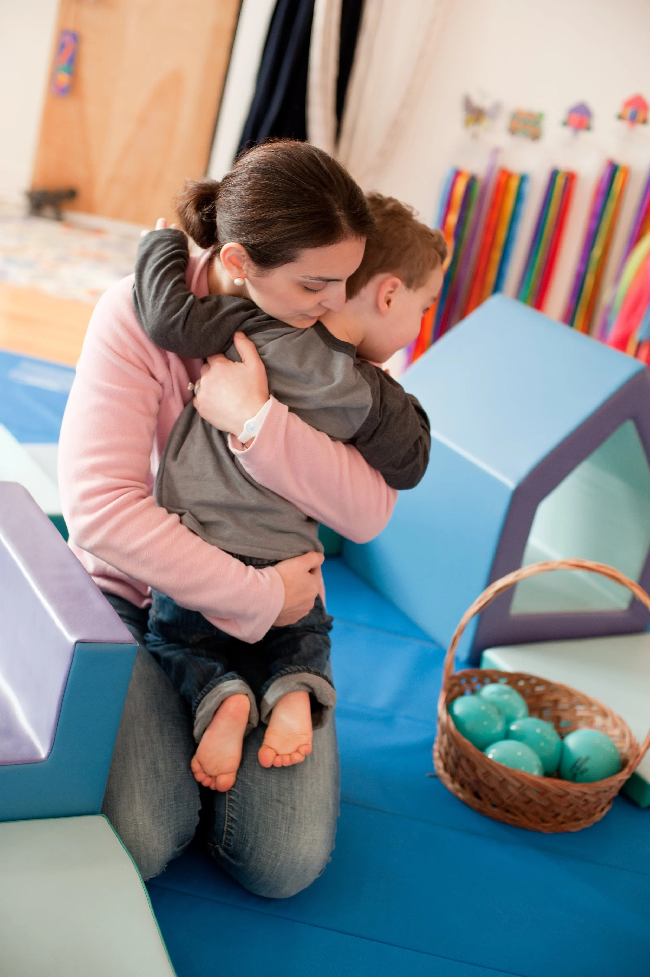 Woman hugging a young boy in a colorful playroom with a basket of plastic eggs nearby.