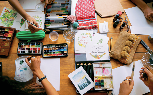 People's hands creating watercolor art on a wooden table with paints, brushes, and art supplies.