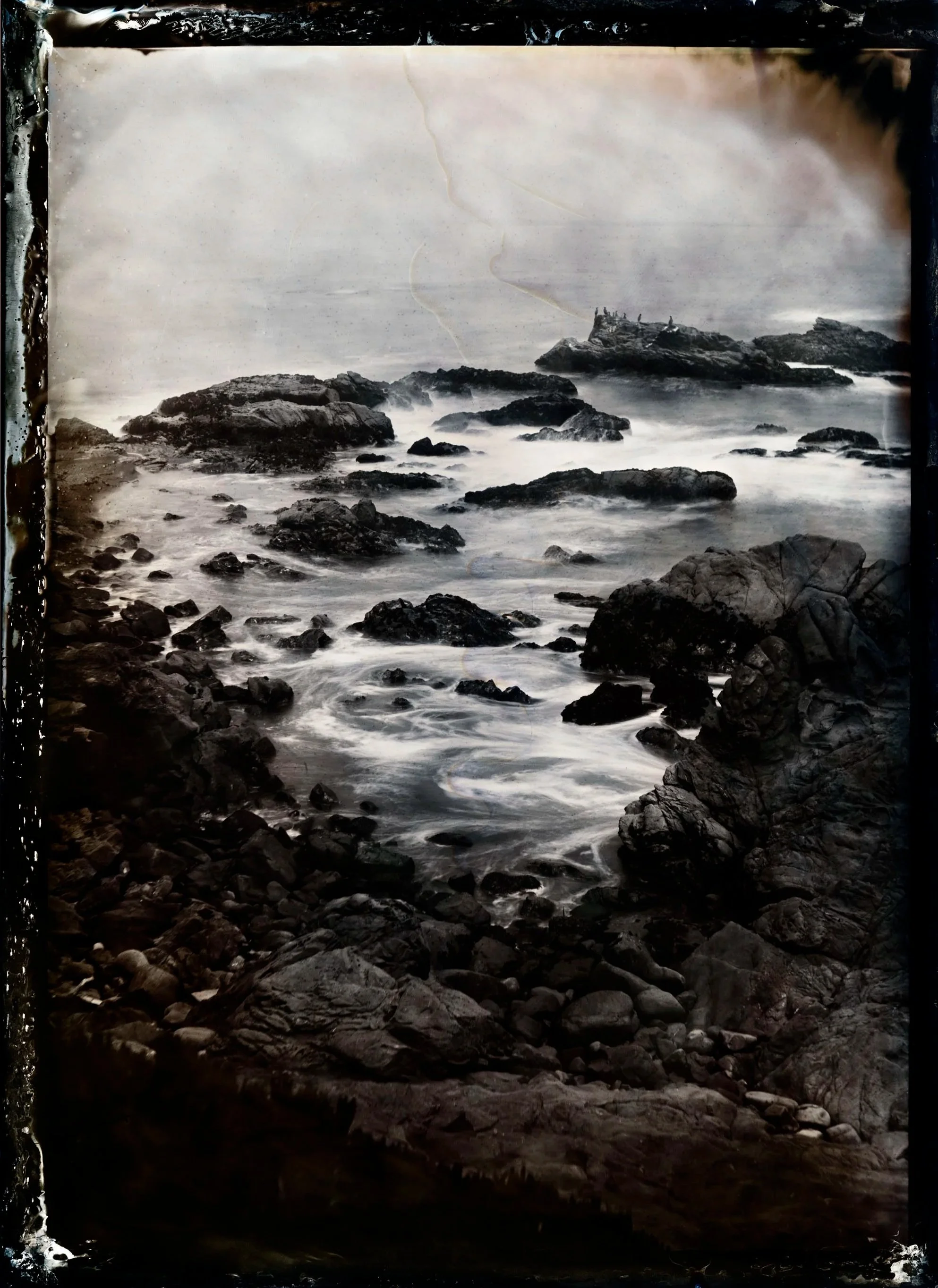 Black and white photograph of a rocky seashore with waves crashing against the rocks and a cloudy sky.