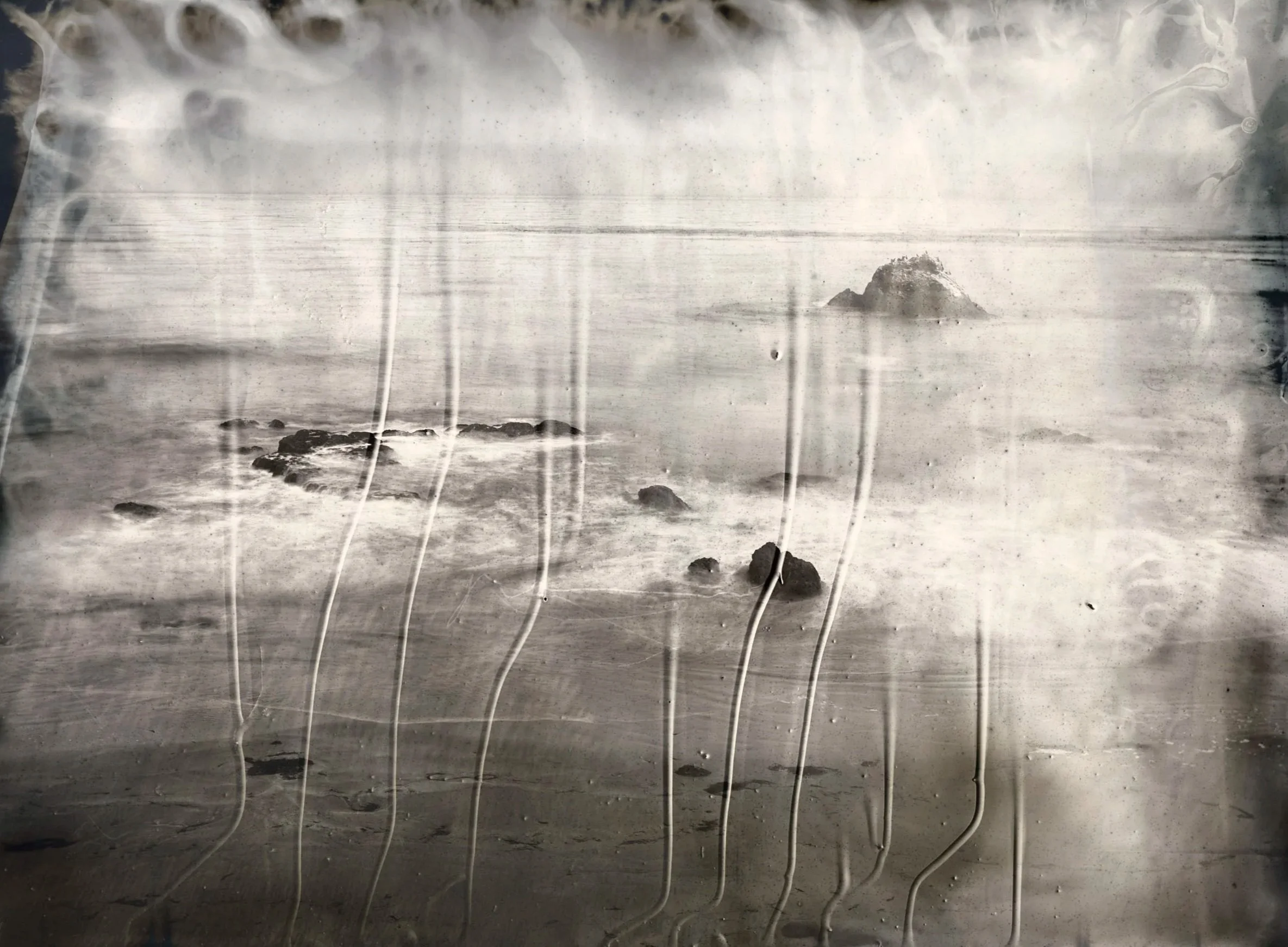 A black and white beach scene with rocks in the water and a small rocky island in the distance, viewed through a scratched glass.