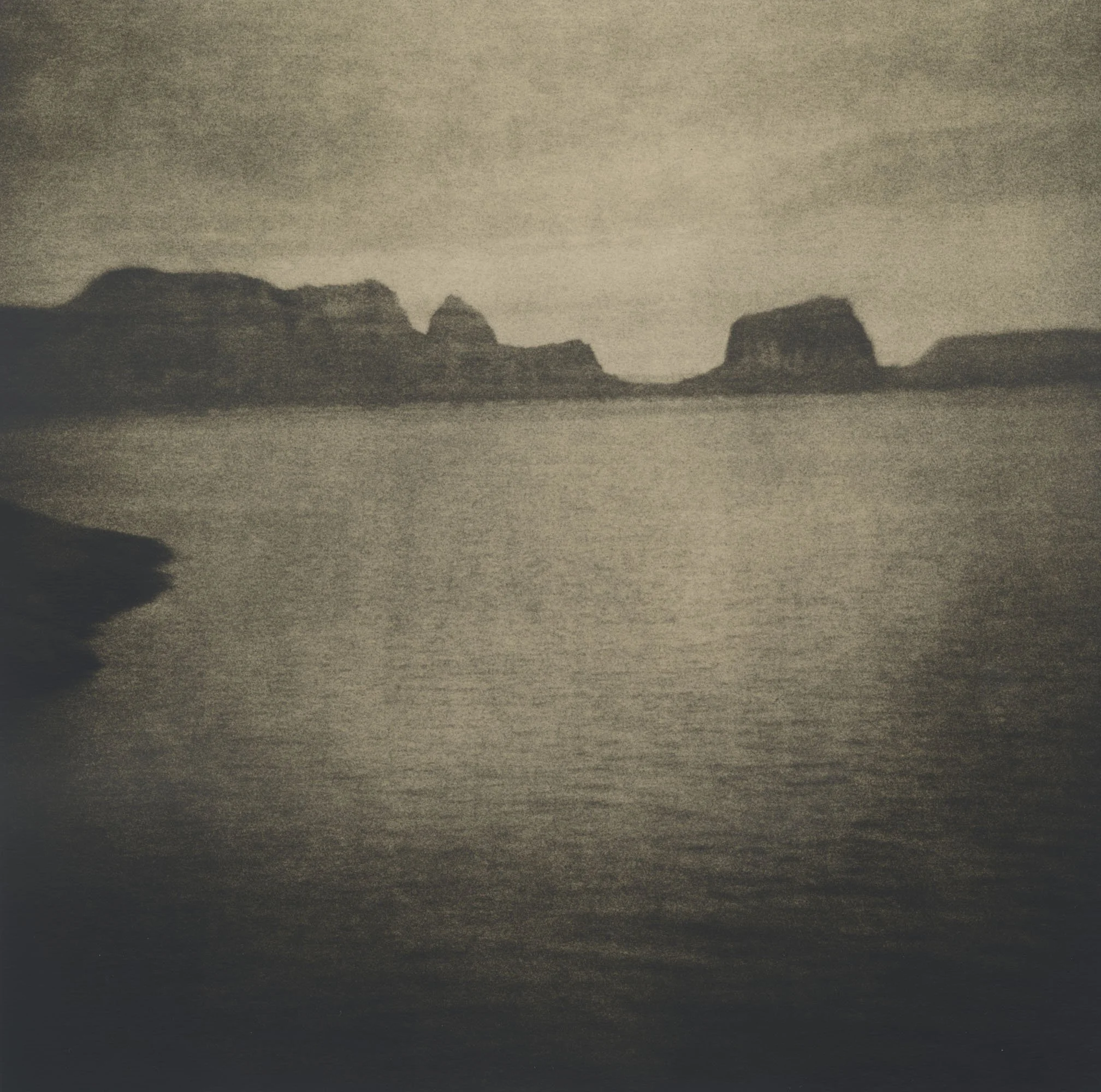 A black and white, grainy photo of a coastline with water in the foreground and rocky cliffs in the background.