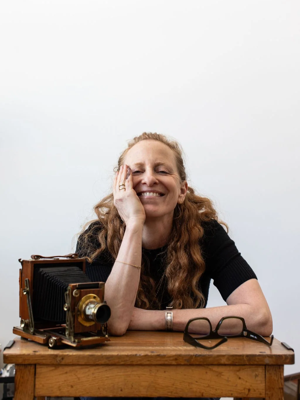 A woman with curly red hair sitting at a wooden table, smiling with her hand resting on her cheek. She is wearing a black shirt, with a vintage large format camera and glasses on the table in front of her.