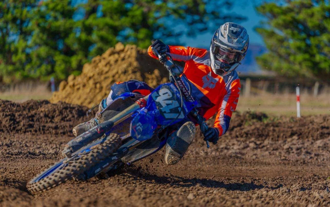 A motocross rider in an orange and blue outfit on a blue dirt bike, racing on a dirt track with trees and a dirt mound in the background.