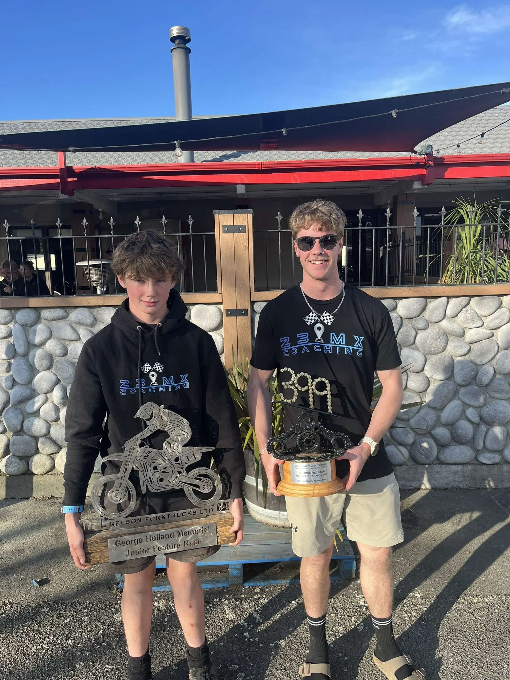 Two young men standing outdoors, holding trophies, wearing black T-shirts with blue text and checkered flags, posing in front of a stone wall and building. The younger holds a motorcycle cutout trophy, while the older has a trophy featuring a gear an