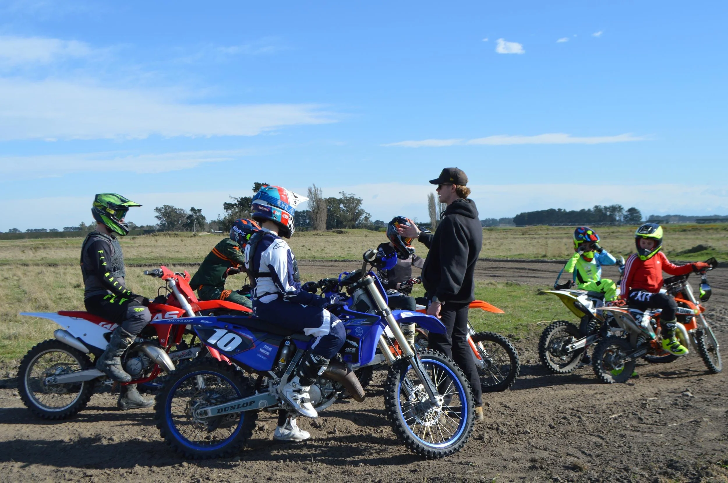 Group of children and a teenager with motocross bikes on a dirt track, receiving instructions from an adult in a black hoodie, under a partly cloudy sky.