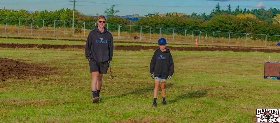 A man and a boy standing on a grassy field. The man is wearing sunglasses, a black hoodie, and shorts. The boy is wearing a blue cap, a black hoodie, and shorts. They are outdoors with a fence and trees in the background.